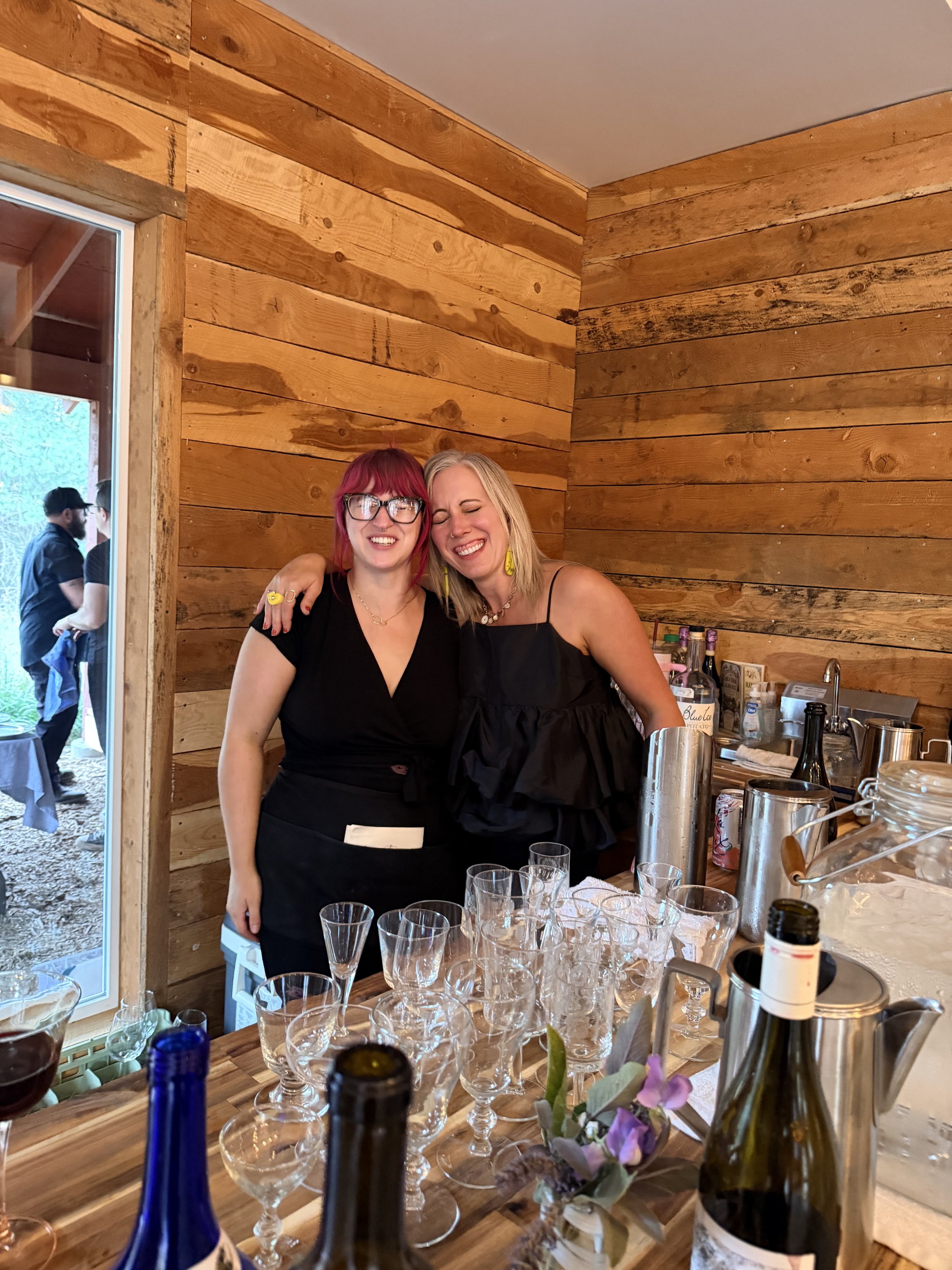 Two women smiling and posing for a photo at a gathering, with a wooden interior wall behind them. The table in front has various glasses and bottles, and there is a small bottle of wine and a flower arrangement on the table.