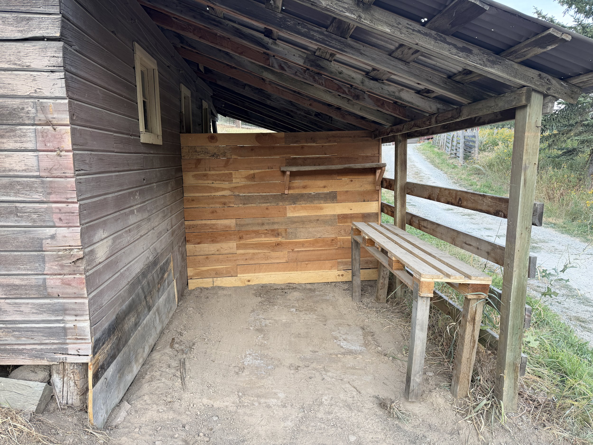 Wooden structure with a slanted roof, a small window, and a bench outside, located along a dirt path with greenery in the background.