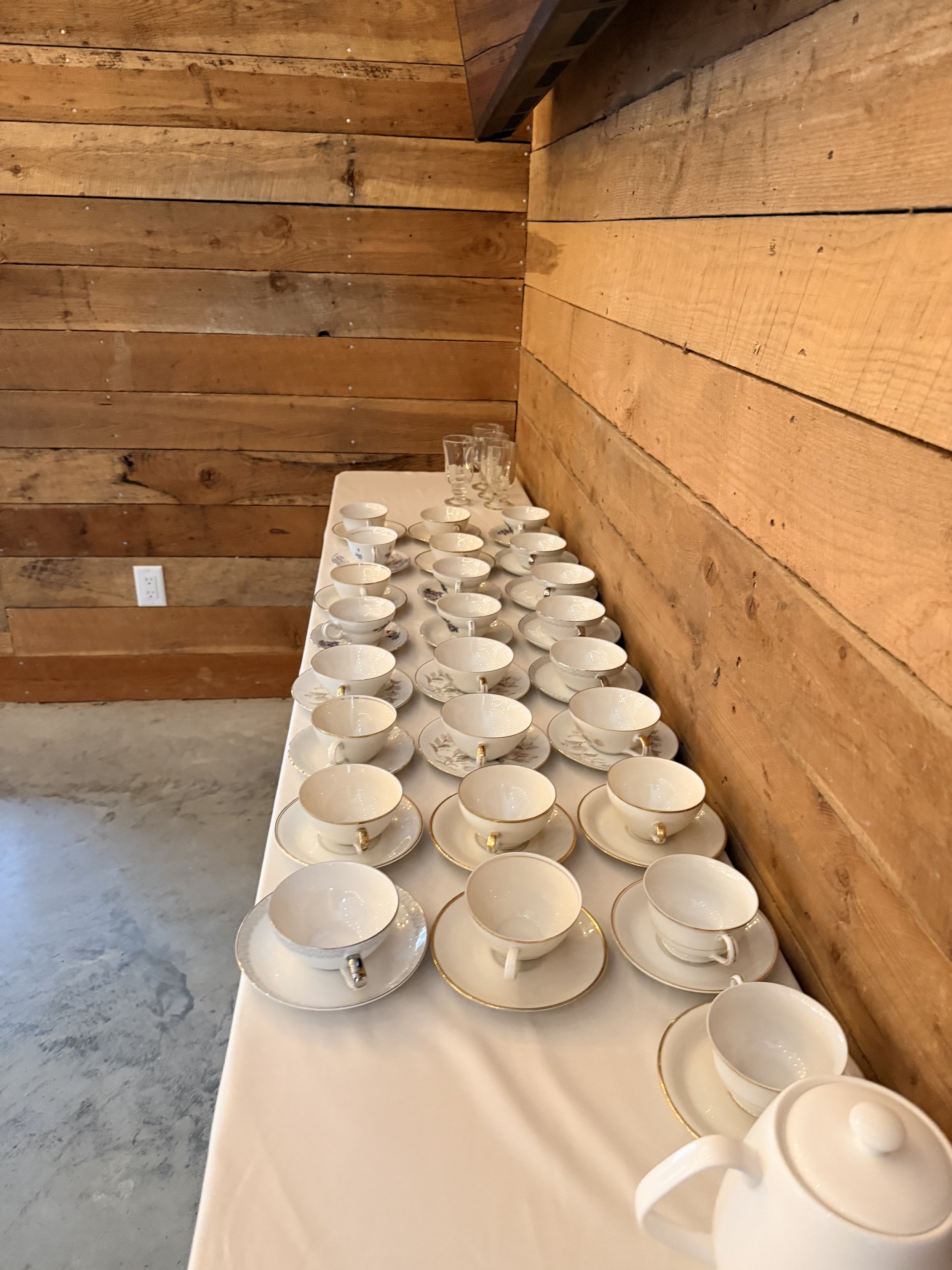 A long table covered with a white tablecloth, set with numerous white teacups and saucers with gold trim, with a wooden wall in the background.