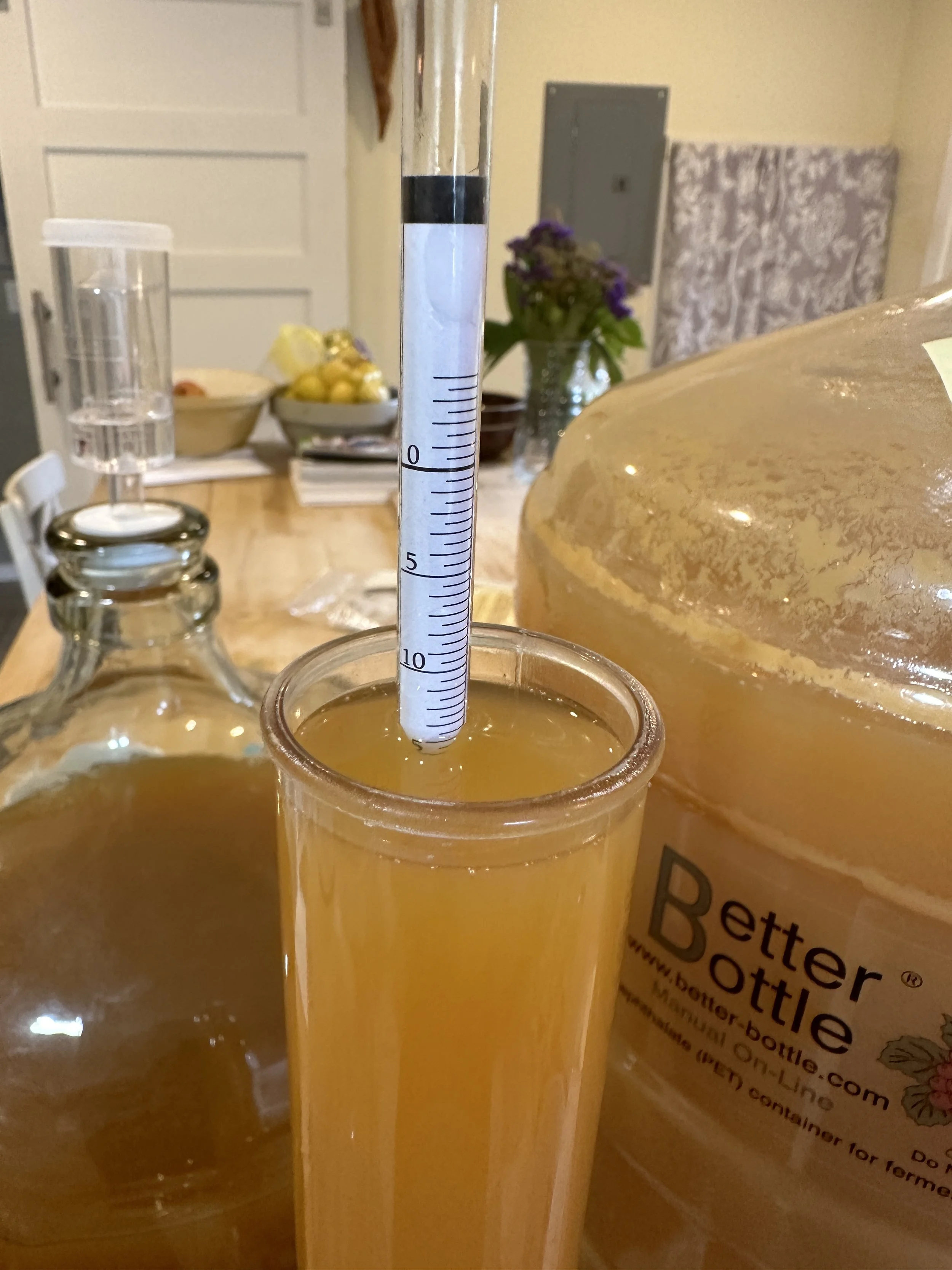 A hydrometer inside a glass of apple juice on a wooden table for testing original gravity and a vase of flowers on a table in a home setting.