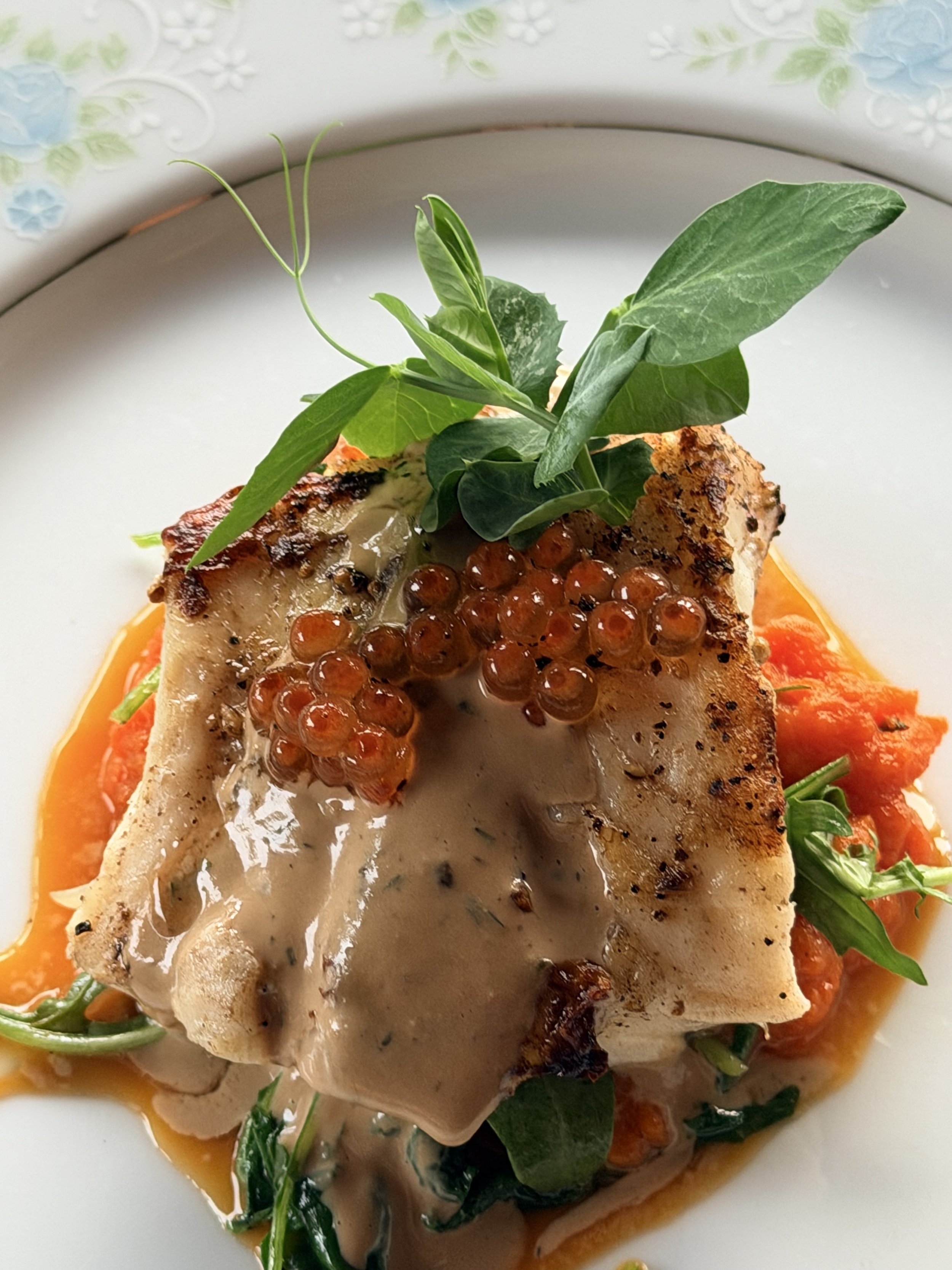 A plated dish featuring a seasoned piece of fish topped with orange caviar, garnished with fresh herbs and greens, served over a bed of cooked vegetables and a creamy sauce.