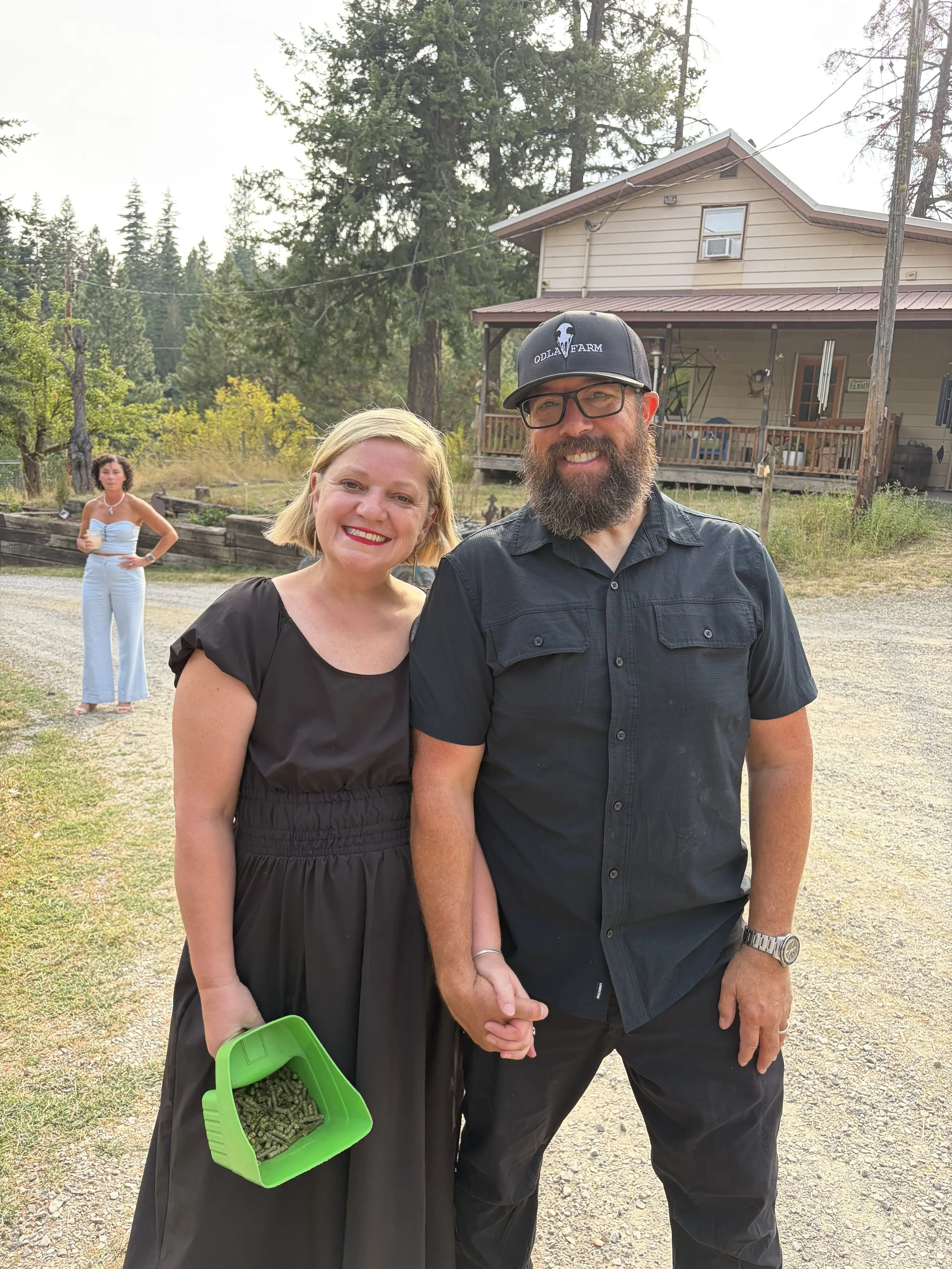 Two people, a woman and a man, standing outdoors and smiling. The woman is holding a green basket filled with alfalfa pellets and the man is in a black shirt and hat, holding hands. There is a house and a woman in the background holding a drink.