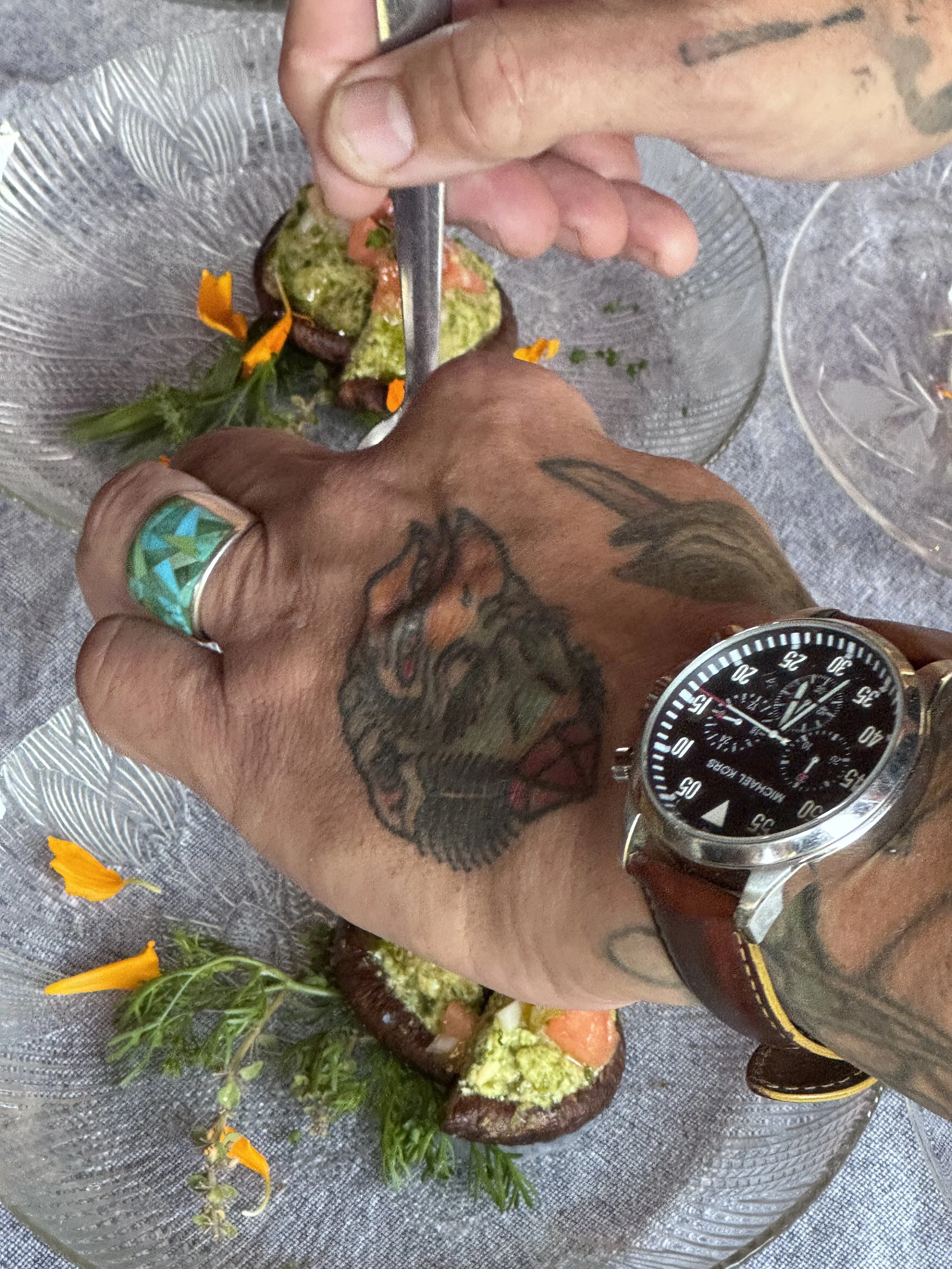 Close-up of a person's hand and wrist with tattoos and a black watch, holding a small metal tool and working on a piece of food on a clear glass plate. The food appears to be an avocado half with various toppings and garnished with orange edible flowers and green herbs.