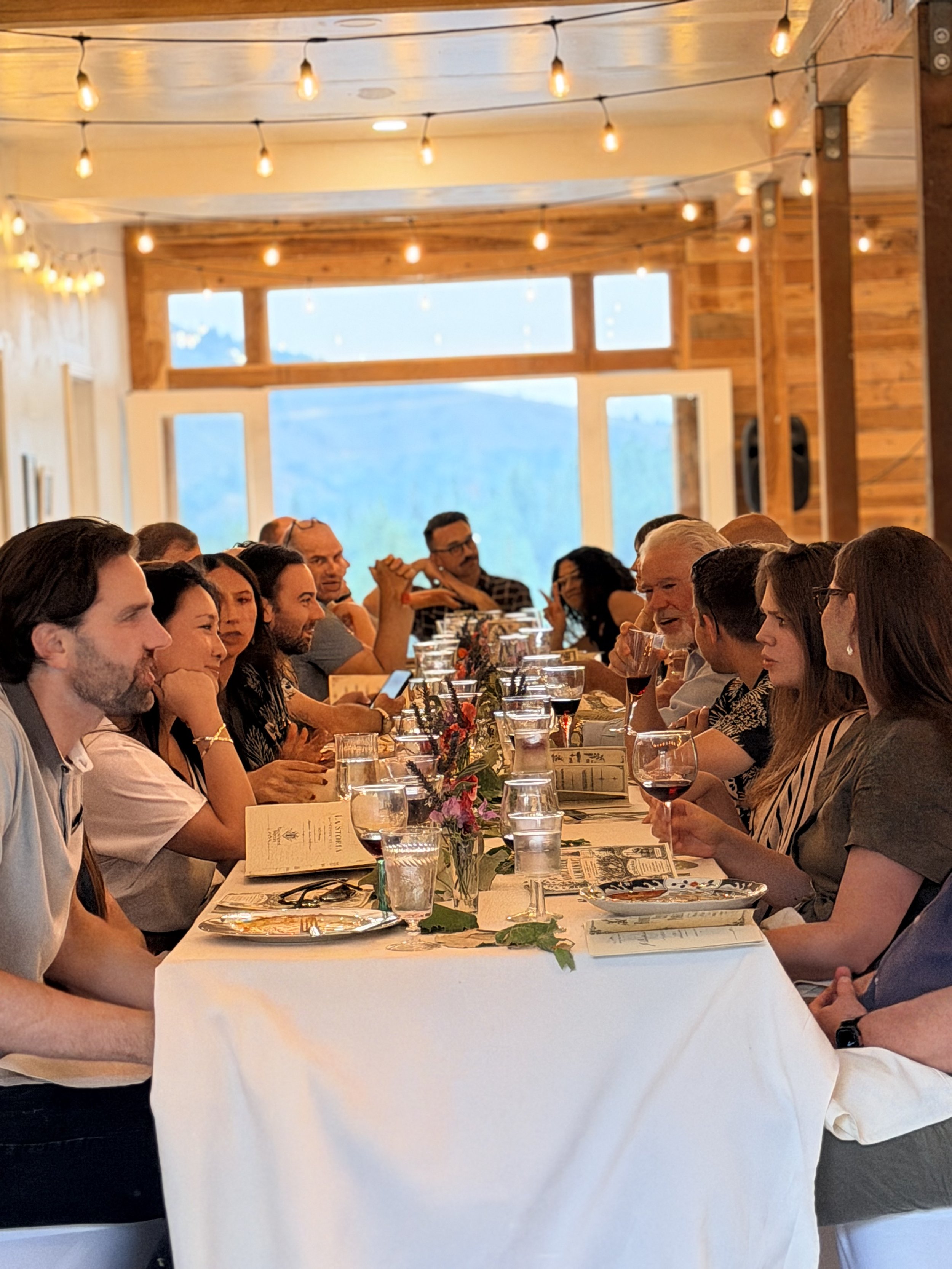 People sitting at a long dining table during a gathering in a rustic wooden venue with string lights and large windows showing a mountain landscape.