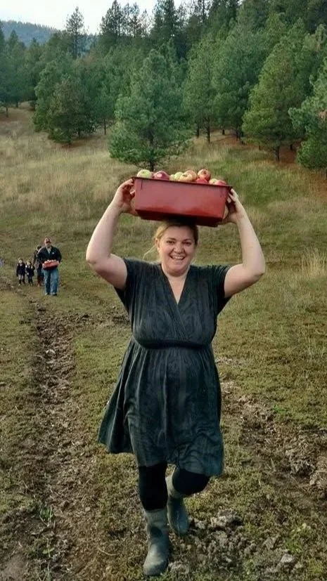 Woman smiling and carrying a red container filled with apples on her head while walking through a farm field, with a group of people in the background and surrounded by lush green trees.