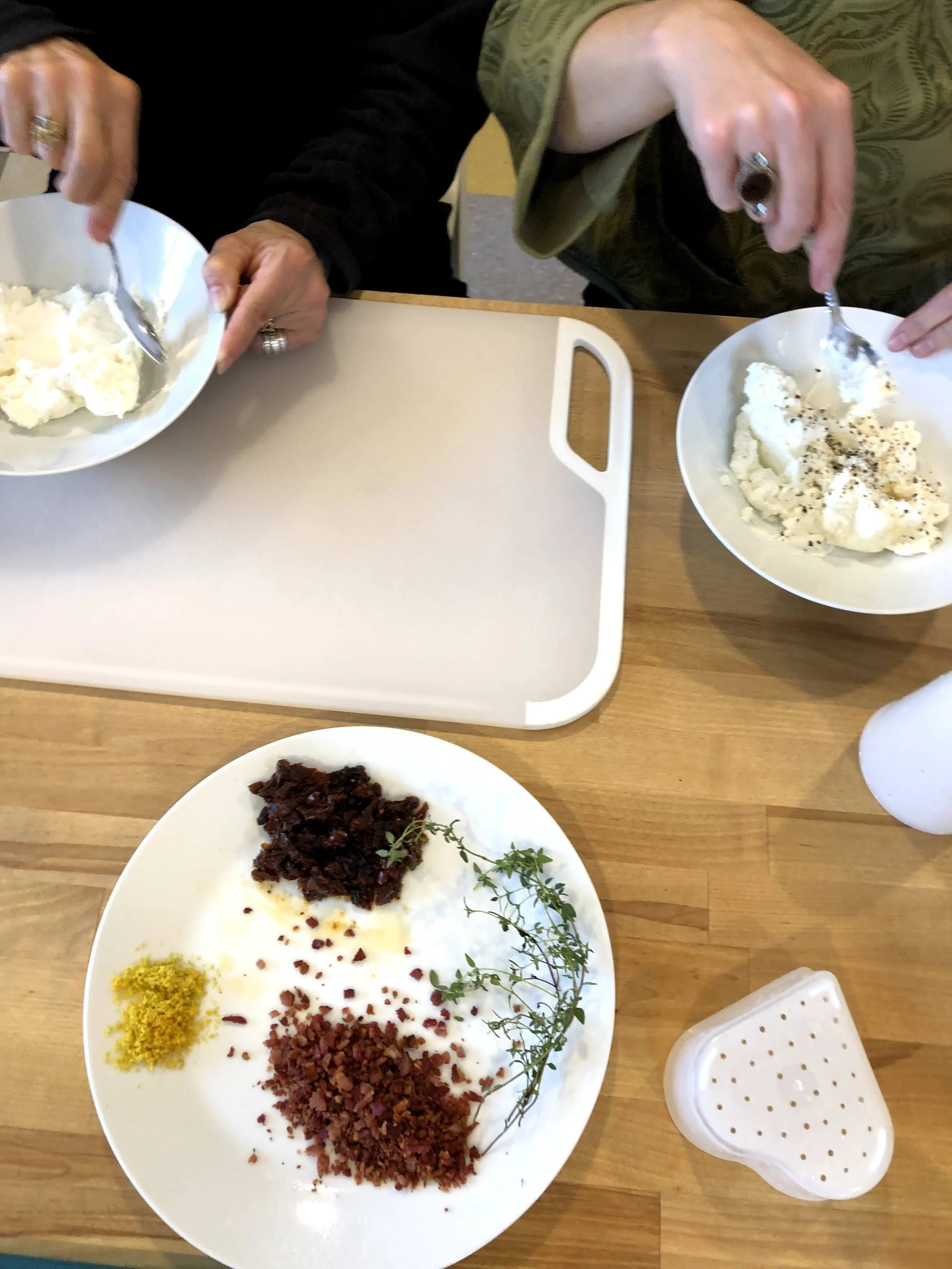 Close-up of a table with three dishes, including a white plate with various colorful spices and herbs, and two white bowls with food being served by two people with their hands. One person is wearing a patterned green shirt, and the other is wearing a black sleeve. The dishes and hands are on a wooden table with a plastic tray nearby.