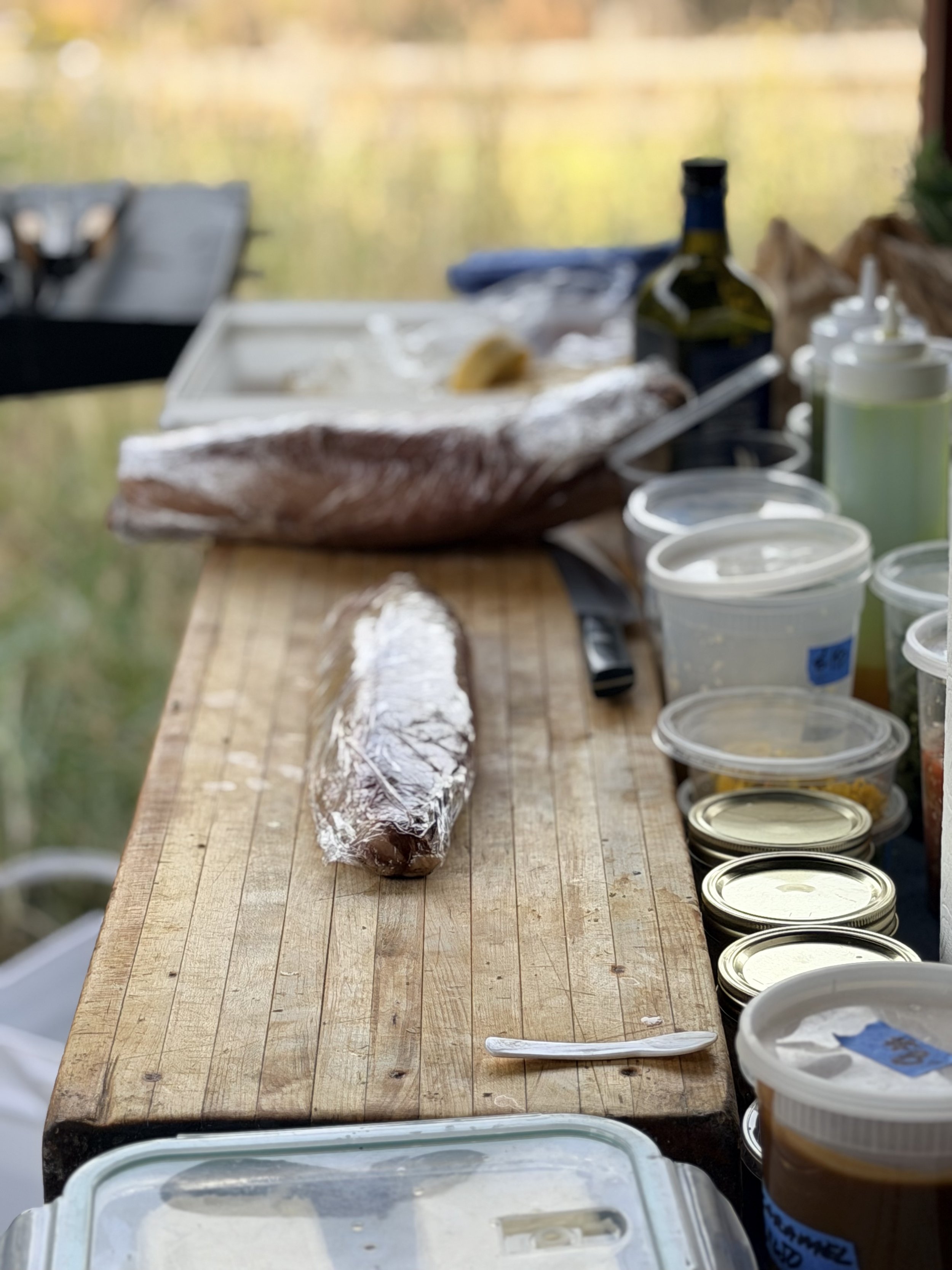 Outdoor scene with a wooden table holding packaged food items, cans, containers with lids, and a bottle, with a blurred background of trees and water.
