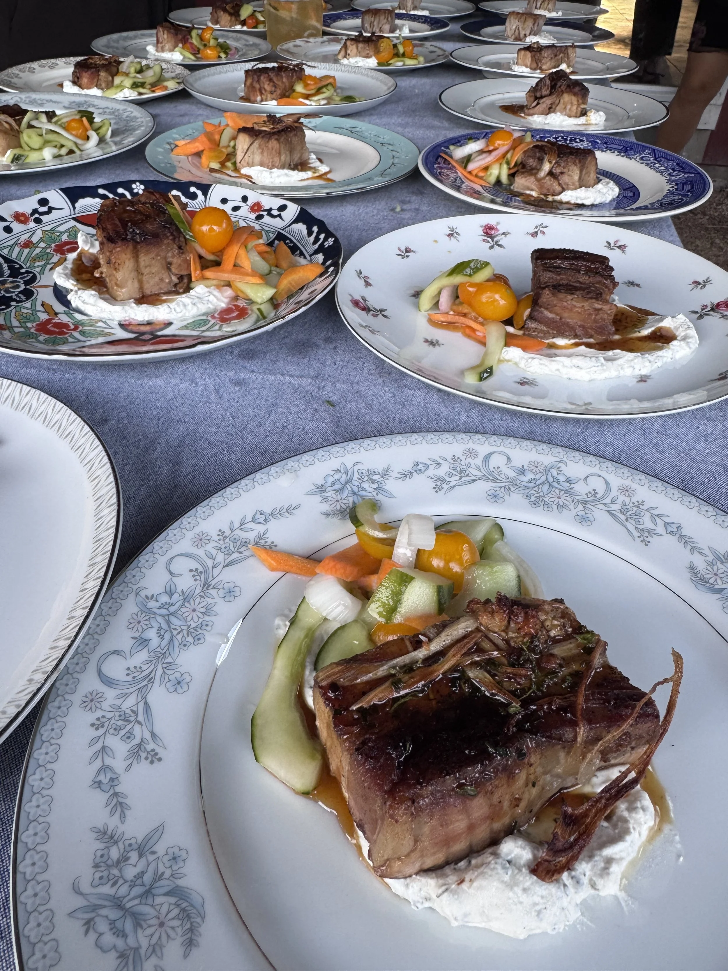 A plated dish featuring a large cut of grilled meat topped with sauce and garnished with vegetables, served on a bed of cream or sauce on a decorative plate.