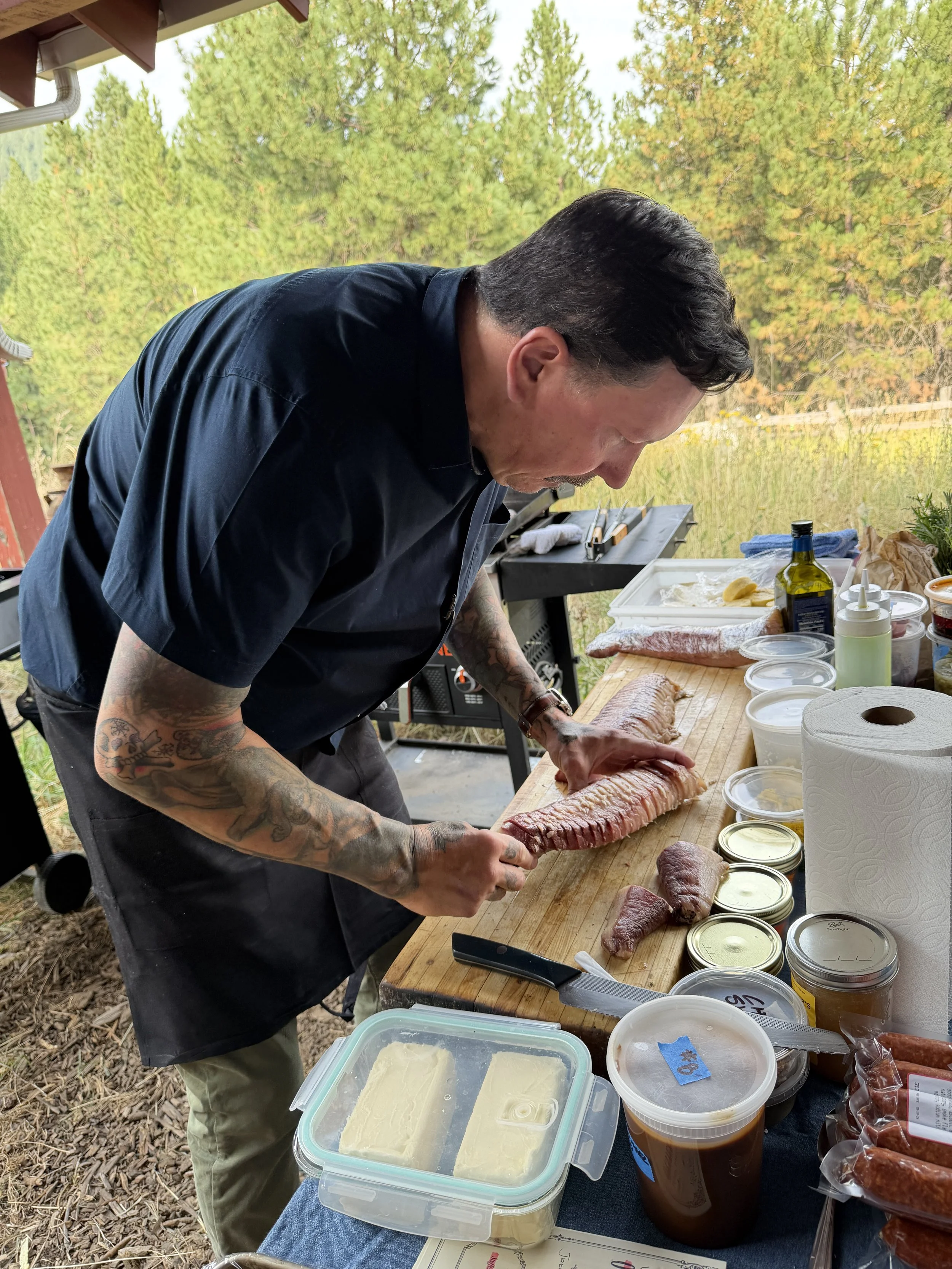 Man preparing fish fillets on an outdoor wooden table with various ingredients, containers, and kitchen tools, surrounded by green trees.