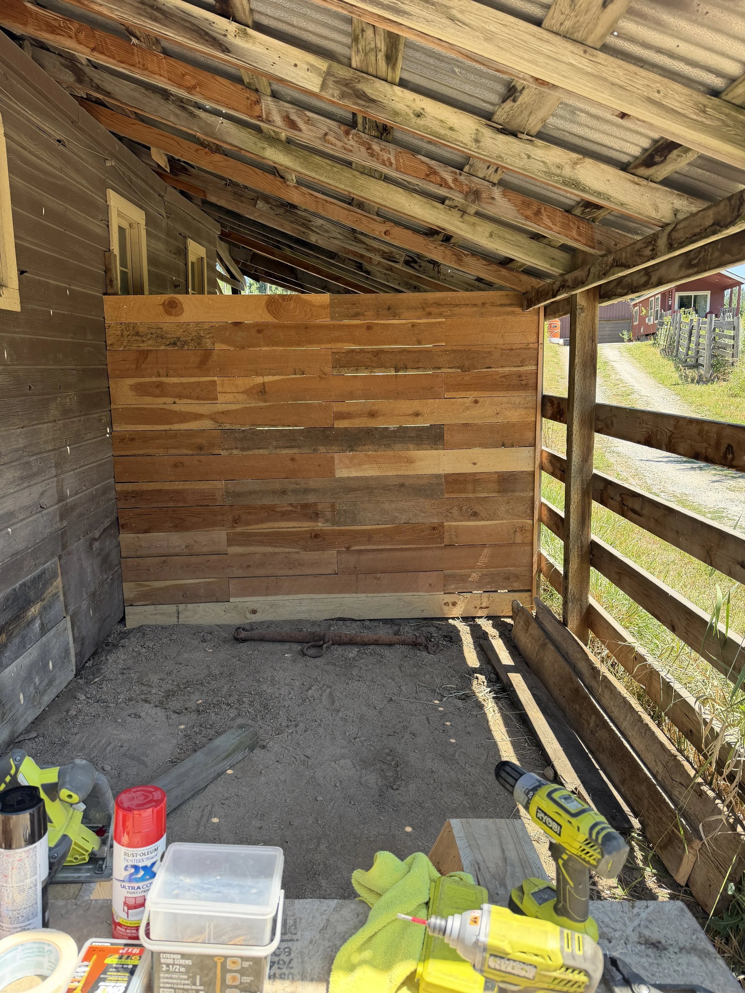 Partially constructed outdoor wooden structure with tools and supplies on a table, including a yellow cordless drill, paint, and miscellaneous construction items, with a dirt ground and surrounding fencing.