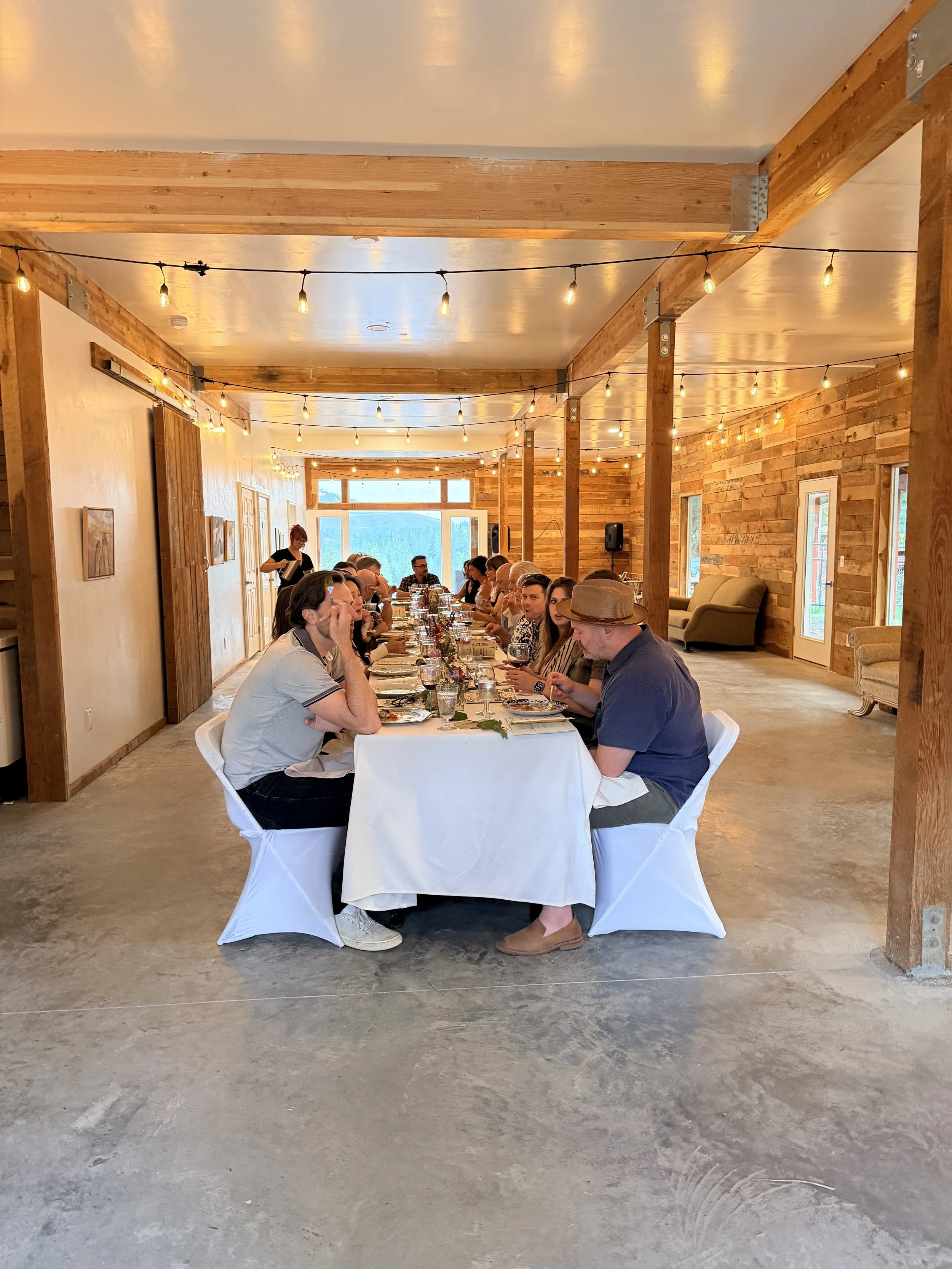 Long rectangular table set for a meal in a rustic wooden hall with string lights overhead and a large window at the back. Several people are seated around the table, engaged in conversation and enjoying the event.
