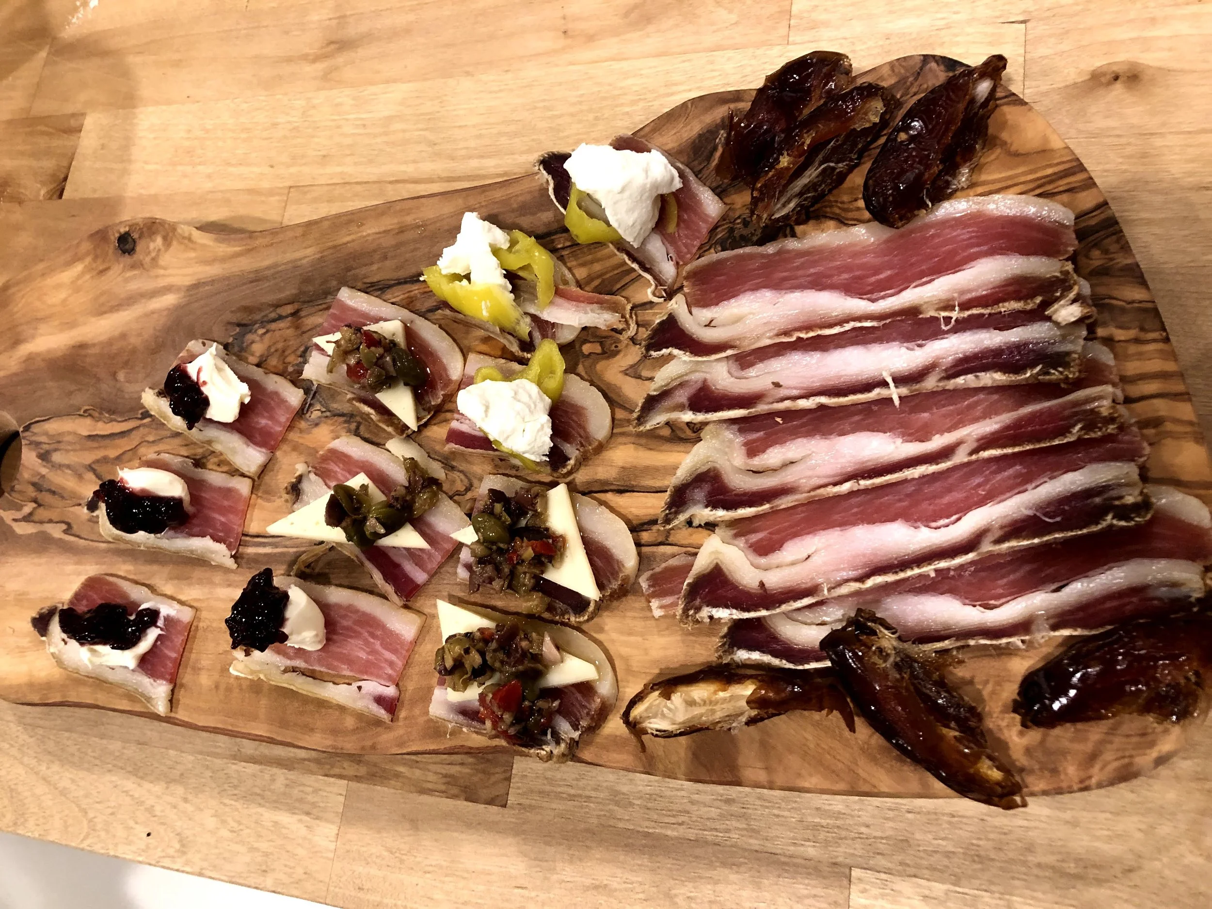 A wooden serving board with slices of cured meats, cheese, and garnishes such as pickles, herbs, and dollops of cream, placed on a wooden table.