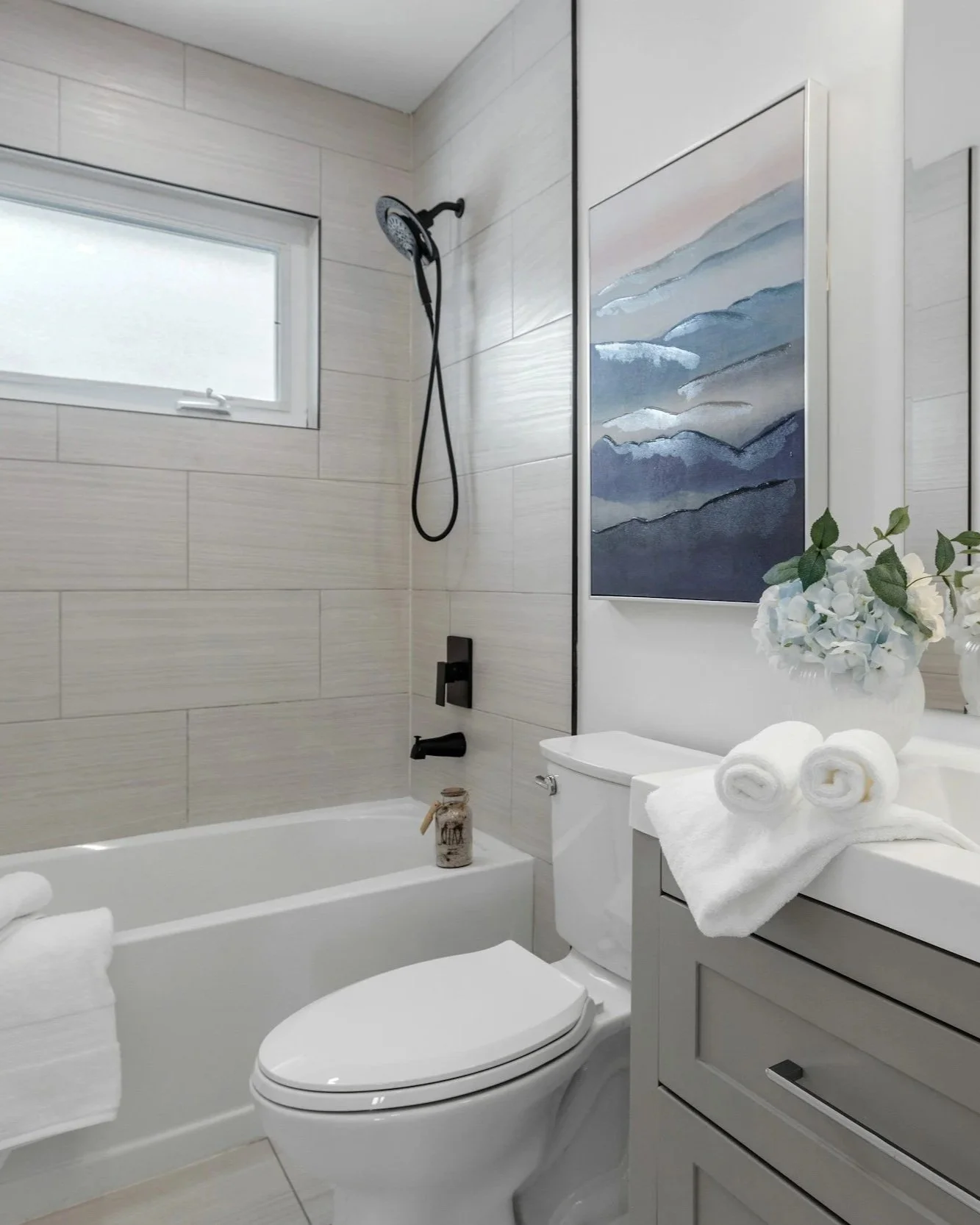 A modern bathroom with a bathtub, toilet, and sink, decorated with rolled white towels, a vase of white flowers, a piece of abstract blue wall art, and a small jar. The bathroom has beige tiled walls, a frosted window, and black fixtures.