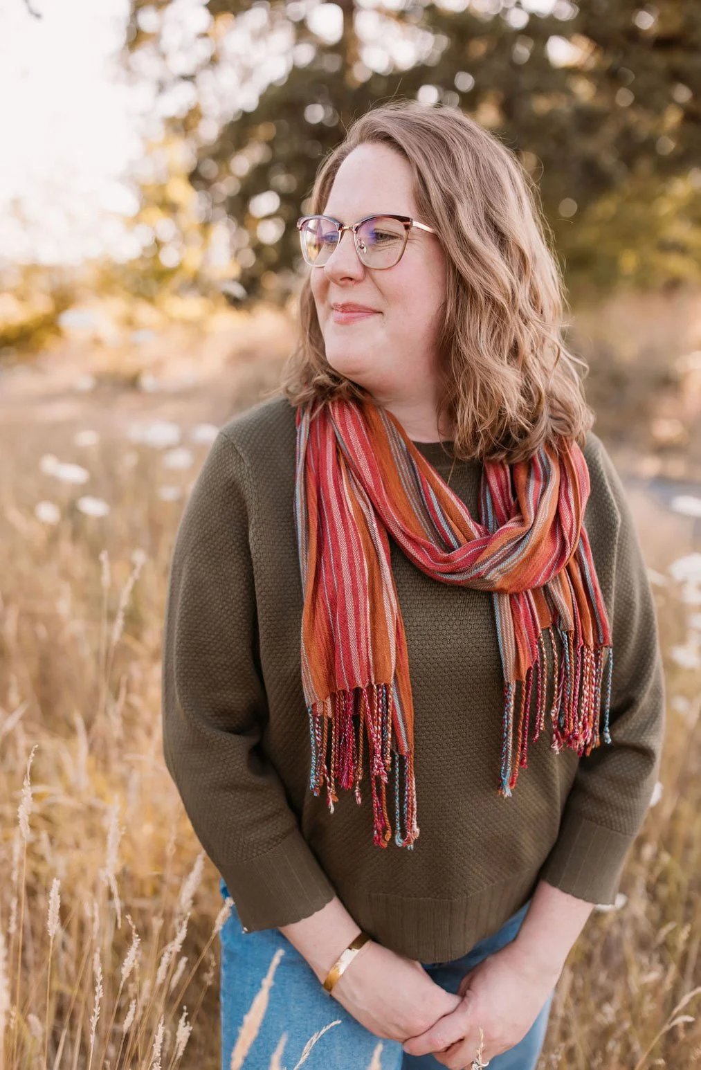 A woman with wavy, shoulder-length hair and glasses stands outdoors in a field of tall, golden grass. She is wearing a green sweater, a colorful striped scarf, and blue jeans, with a warm, fall background