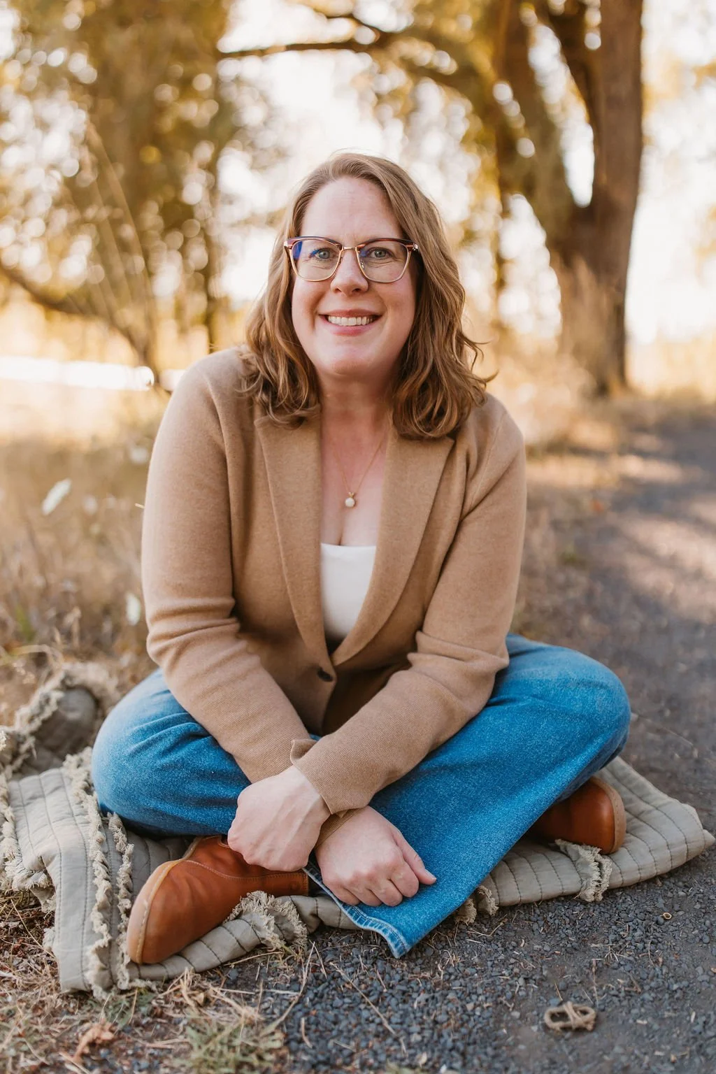 A woman with glasses and wavy brown hair sitting cross-legged on a blanket outdoors surrounded by autumn trees, smiling at the camera.