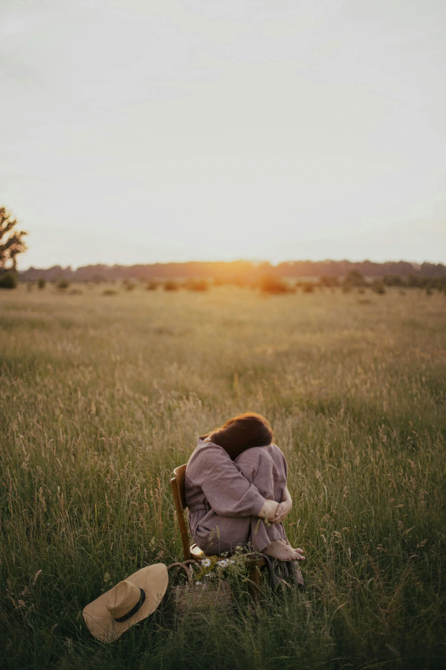 A girl sitting in a grassy field during sunset, hugging her knees with her head down. A straw hat is placed on the ground nearby, and a woven basket with flowers is beside her.