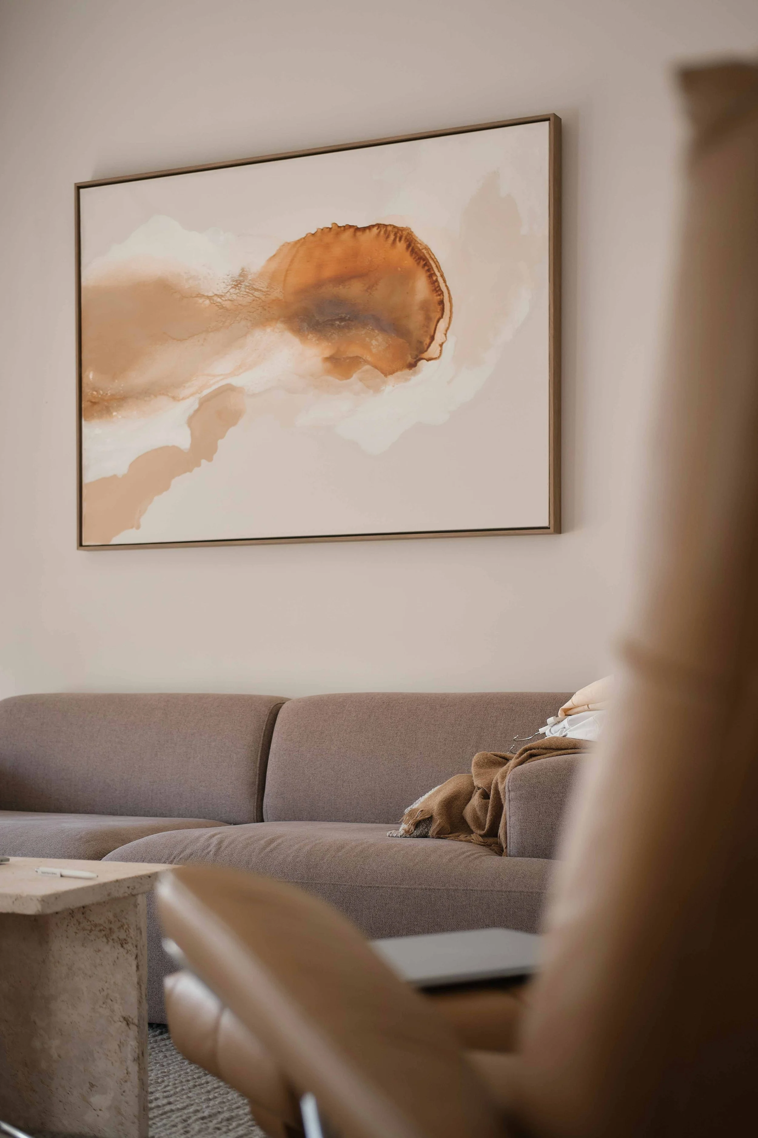 Living room with a beige sofa, abstract artwork on the wall, a marble coffee table, and a person sitting on a chair with a blanket and face mask.