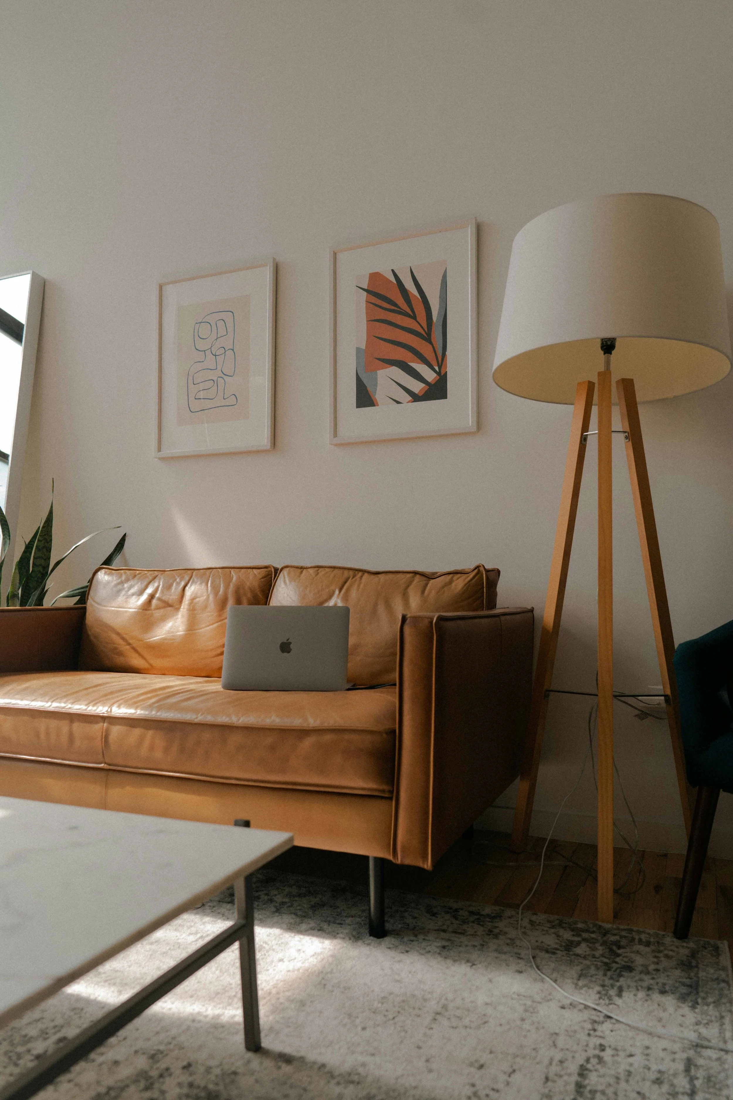 Living room with tan leather sofa, white wall with two framed abstract art prints, wooden tripod floor lamp, and a laptop on the sofa.