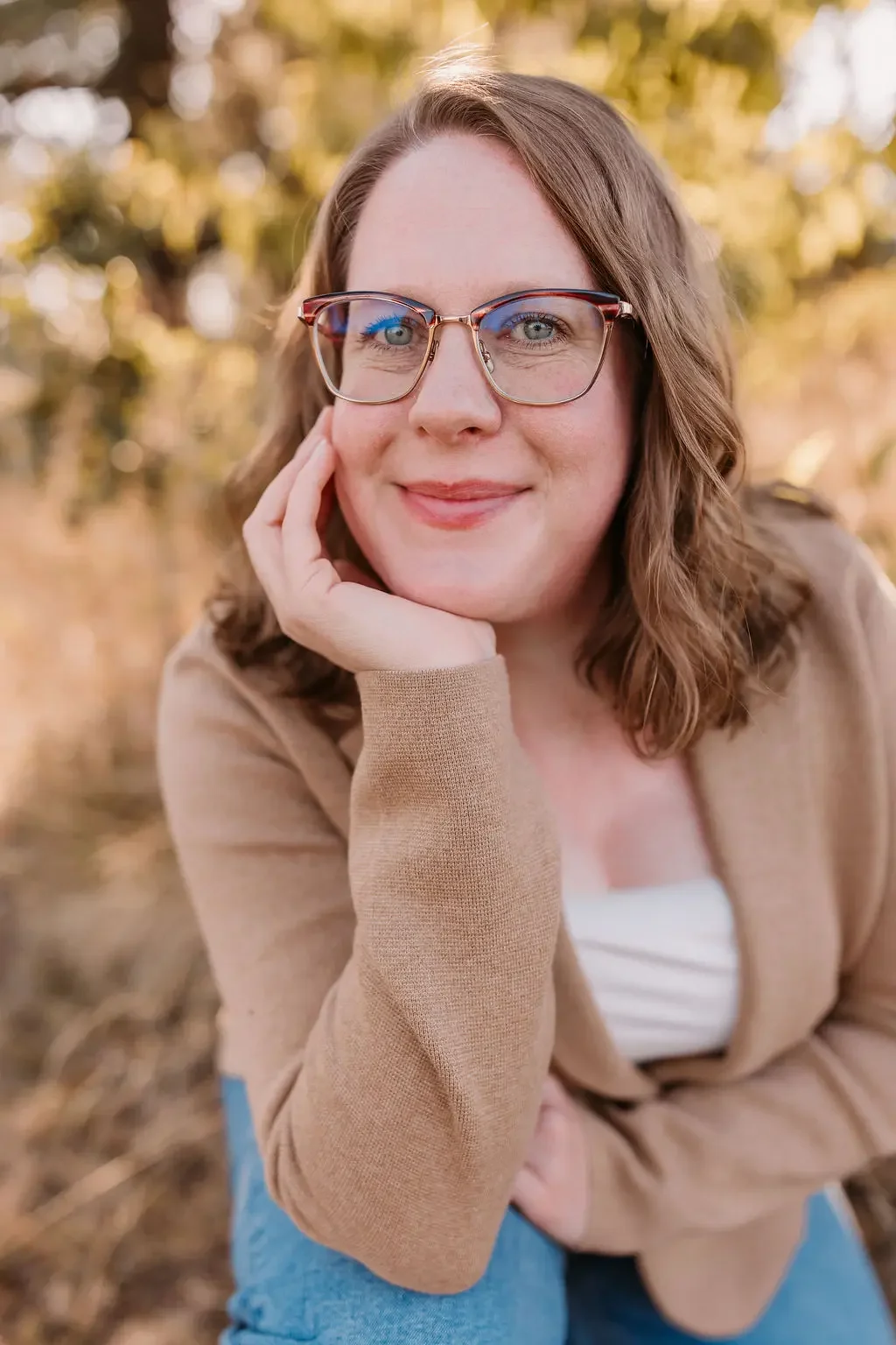 A woman with shoulder-length brown hair and glasses, smiling outdoors in autumn, resting her chin on her hand.