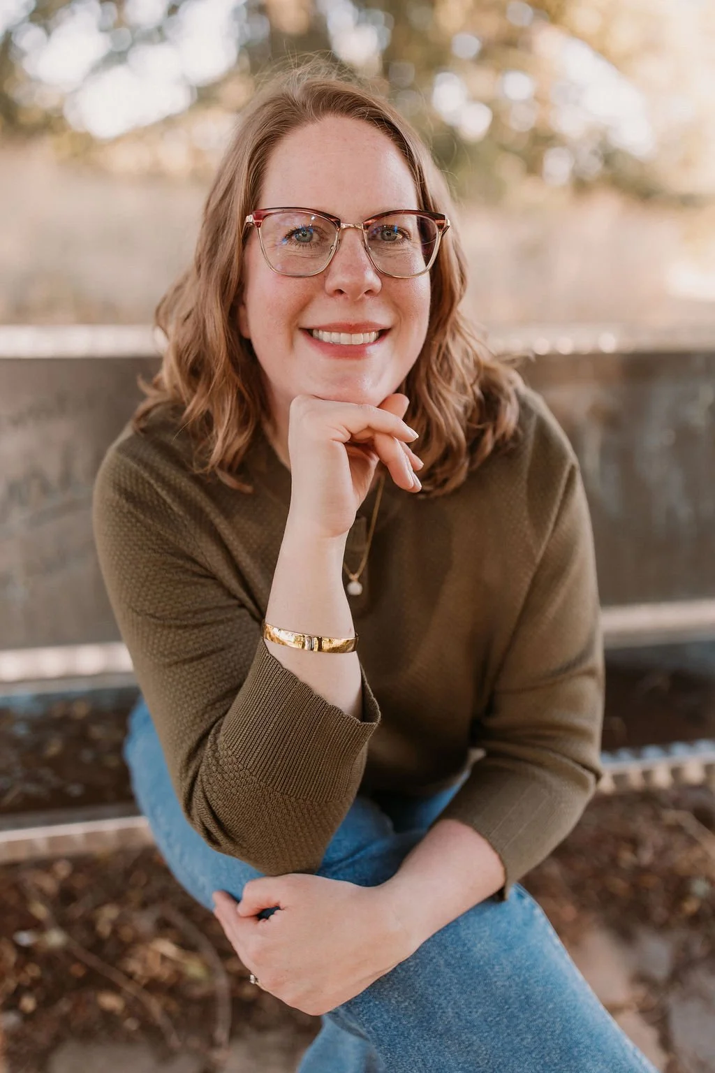 A woman with shoulder-length wavy hair, wearing glasses, a brown sweater, and blue jeans, sitting on a park bench with a blurred natural background, smiling at the camera.