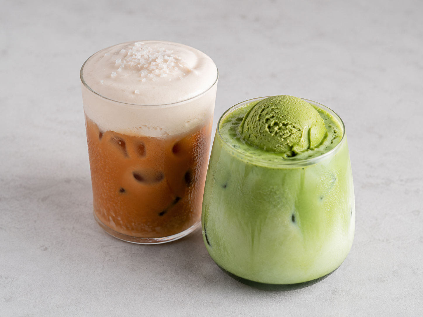 Glass of iced coffee with foam topping and salt, and glass of iced matcha latte topped with matcha ice cream