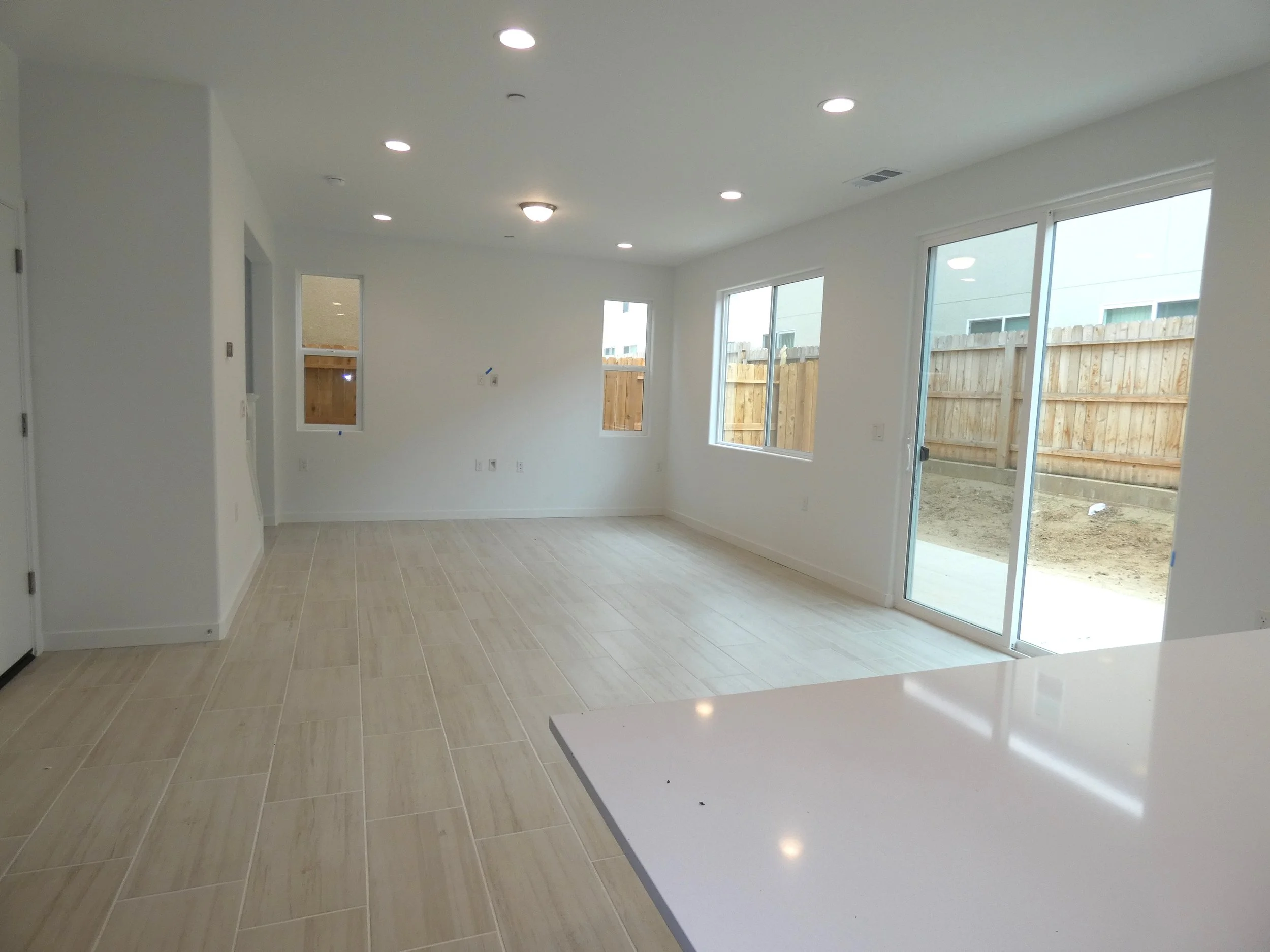 Empty living room with large glass sliding doors leading outside, white walls, beige tile flooring, and recessed ceiling lights.