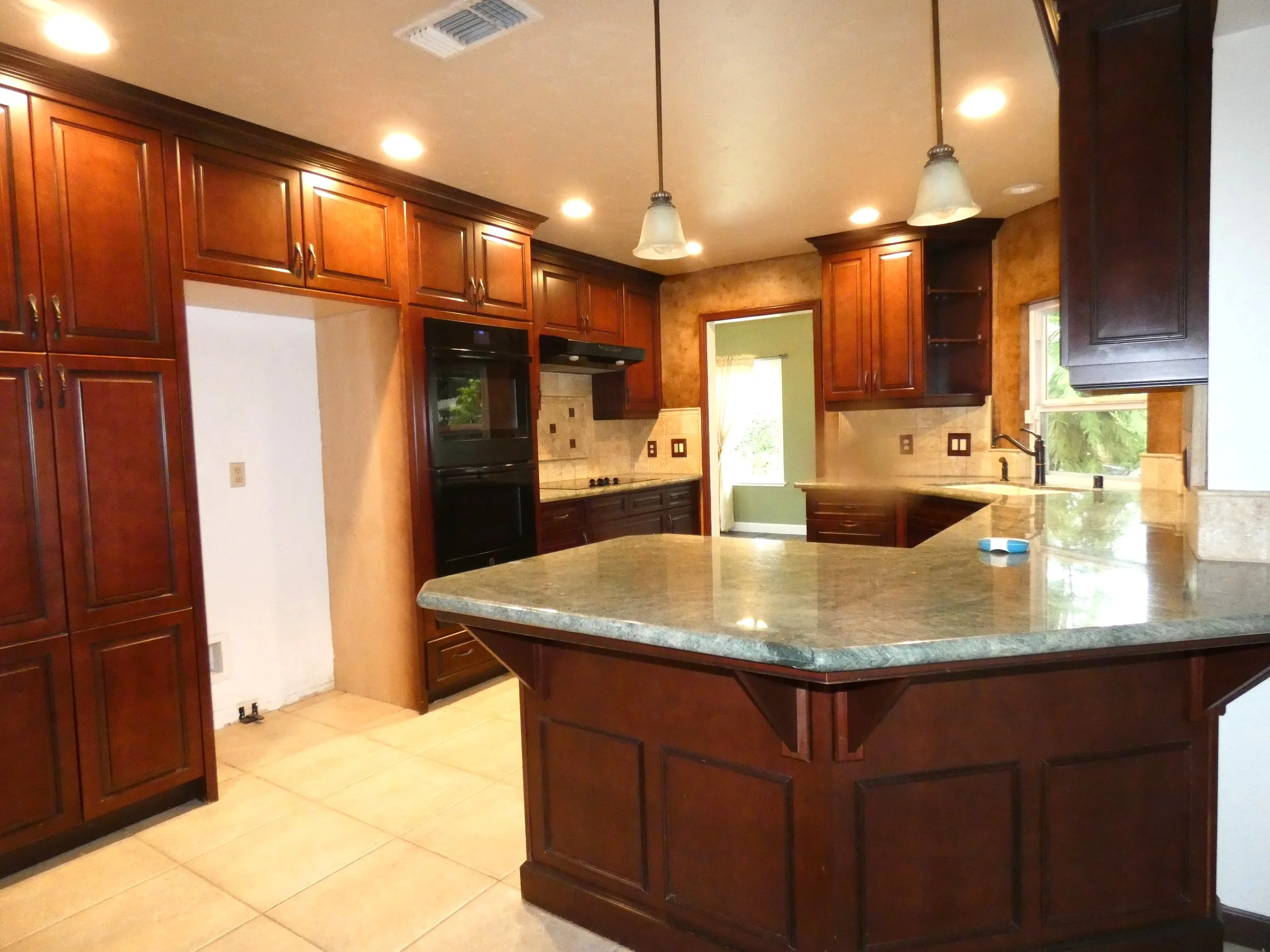Kitchen with dark wood cabinets, beige tiled floor, granite countertop, black appliances, and a window with a view of trees outside.