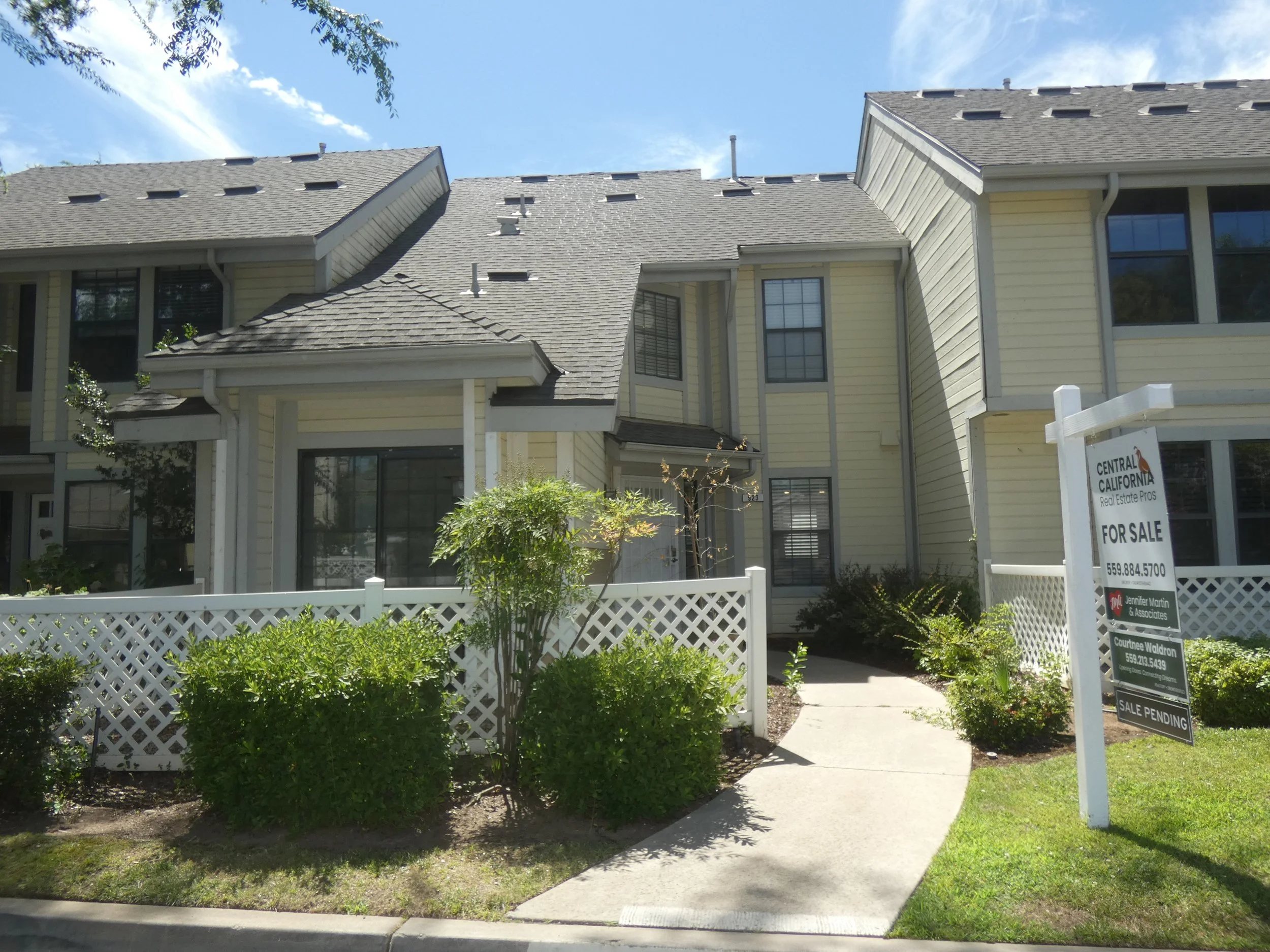 A two-story, yellow house with a grey roof, a white picket fence in front, lush green bushes, and a for sale sign outside.