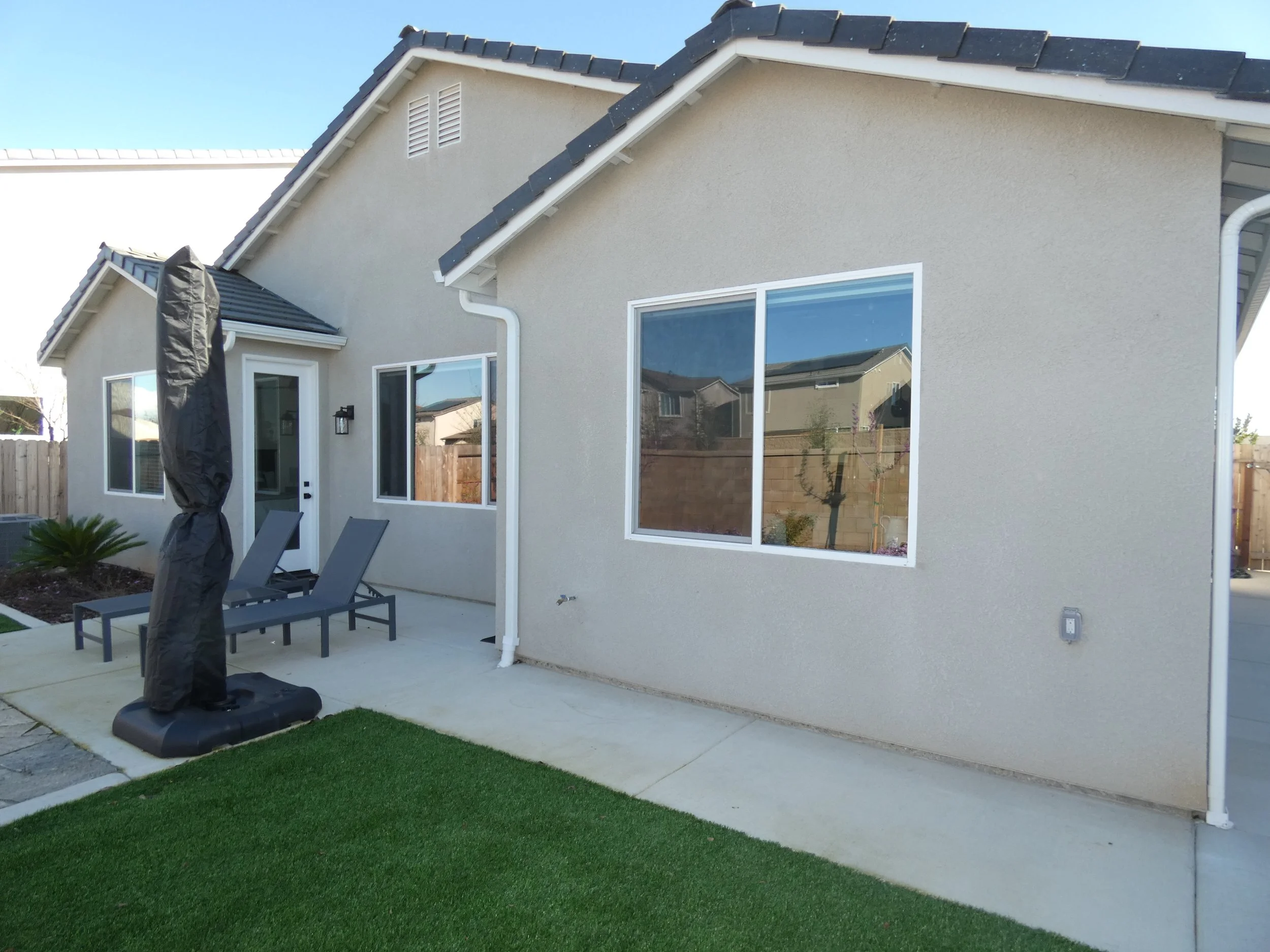 Backyard patio with a concrete slab, two lounge chairs, and a black covered grill. The house has beige stucco walls, large sliding glass doors, and a window. There are kitchen or living room window screens on the house, a small palm plant, and a gree
