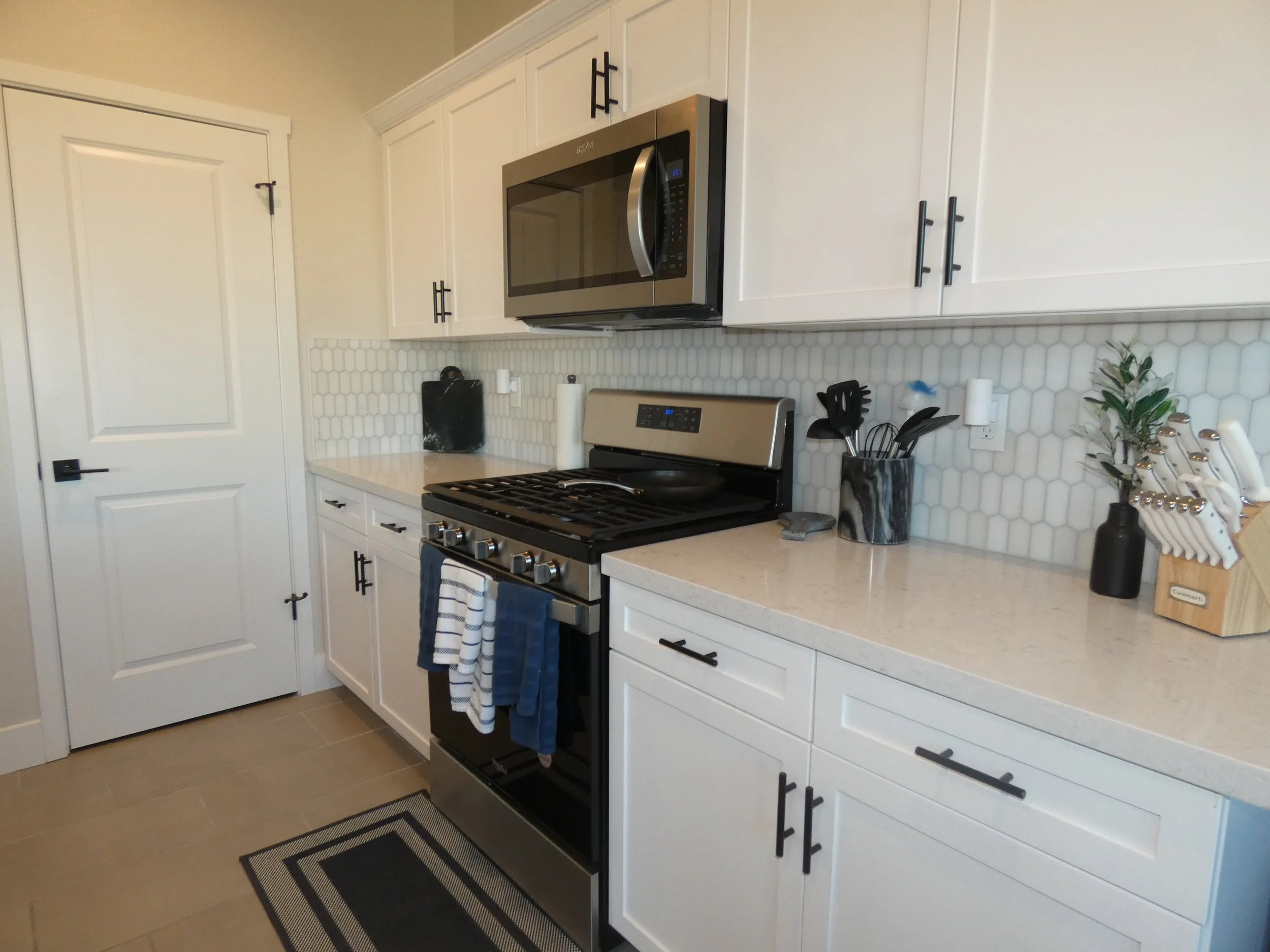 A modern kitchen with white cabinets, a stainless steel microwave, and a black stove with a black and white striped towel hanging on the oven handle. There are kitchen utensils in a marble-patterned container, paper towels, and a green leafy plant on