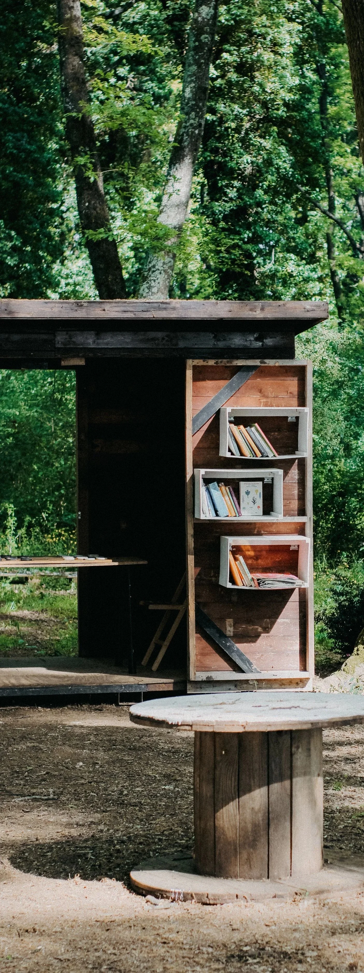 A small wooden outdoor library with multiple shelves filled with books, set against a backdrop of green trees and foliage, with a circular wooden table in the foreground.