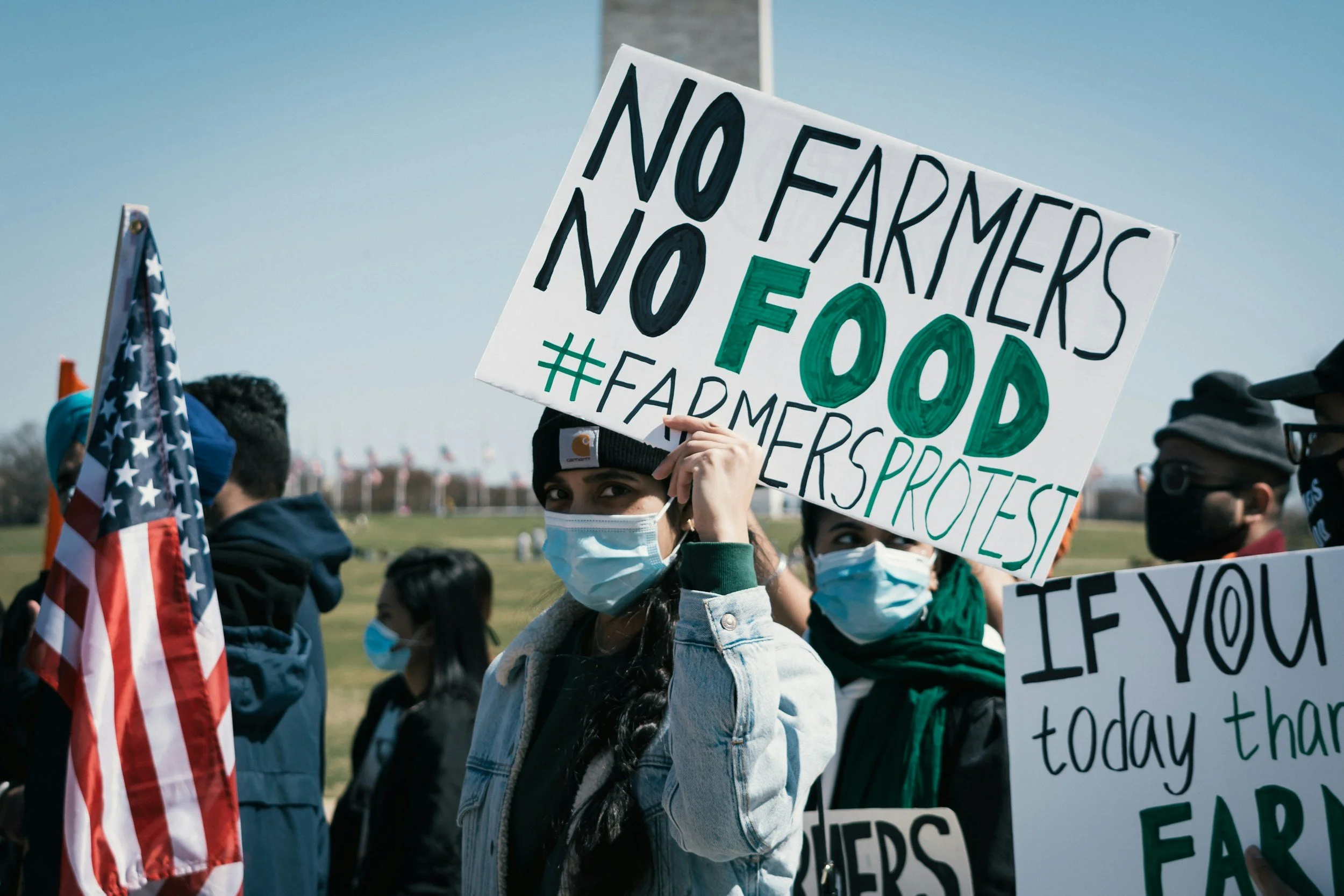 A group of protesters wearing face masks at an outdoor rally. One person holds a sign that reads, 'NO FEAR MOTHERS NO FOOD #FARMERS PROTEST.' An American flag is also visible.