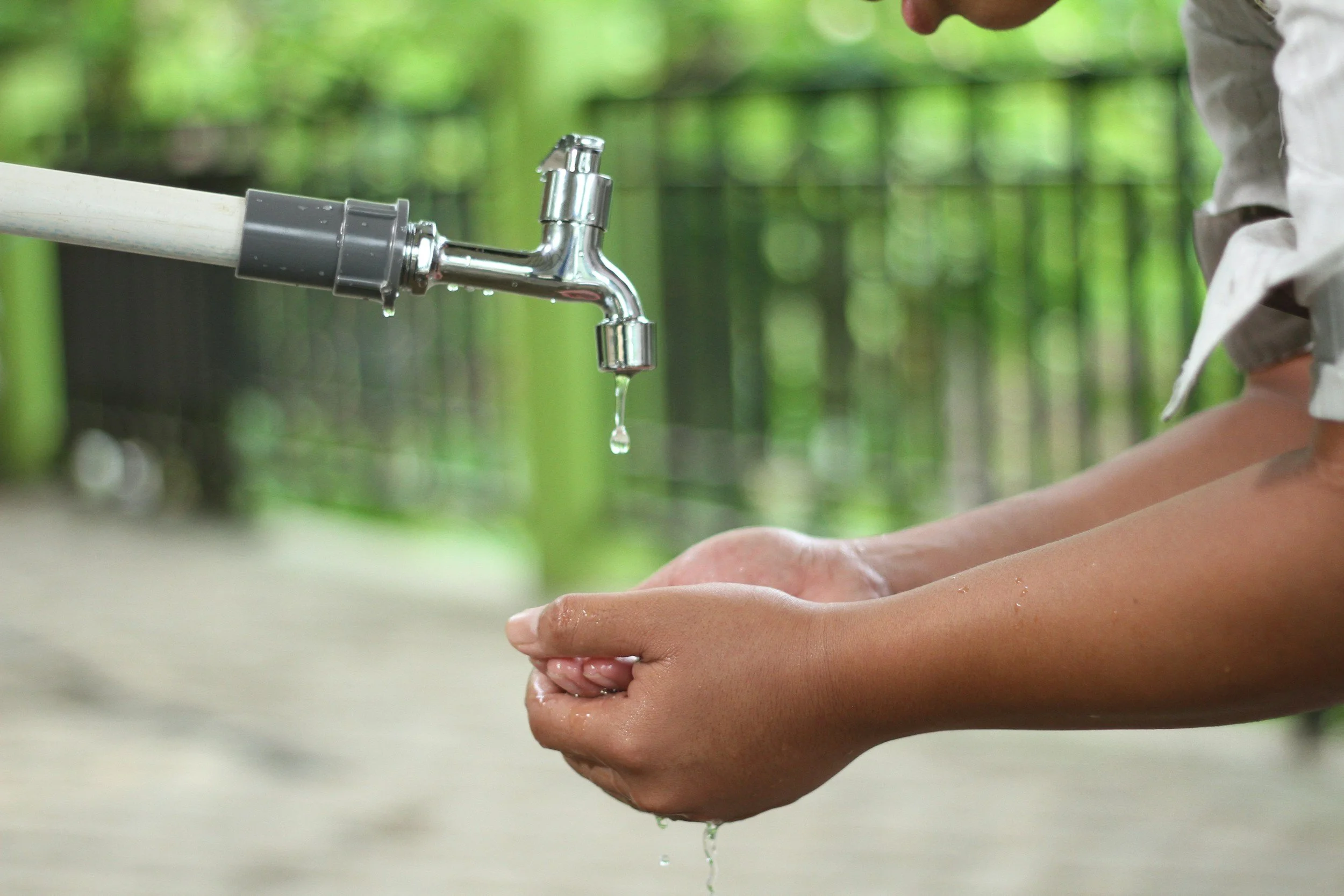 Child washing hands with water from an outdoor faucet in a natural setting.