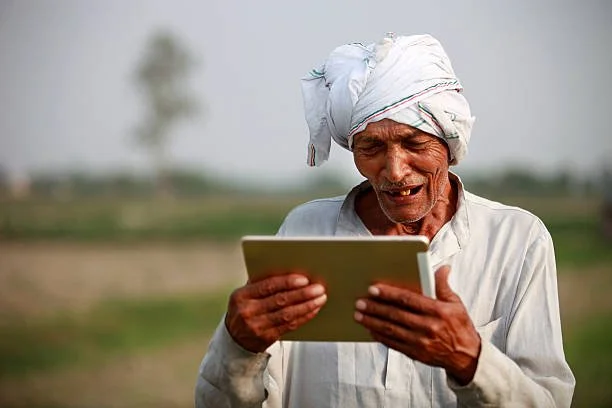 An elderly man with a turban looking at a tablet outdoors in a rural area.