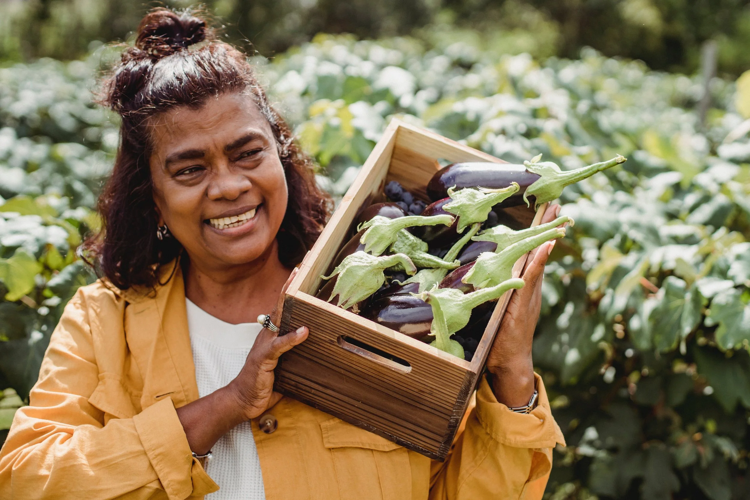 A woman with dark hair in a top bun holding a wooden box filled with eggplants and green stems, standing outdoors among green foliage.