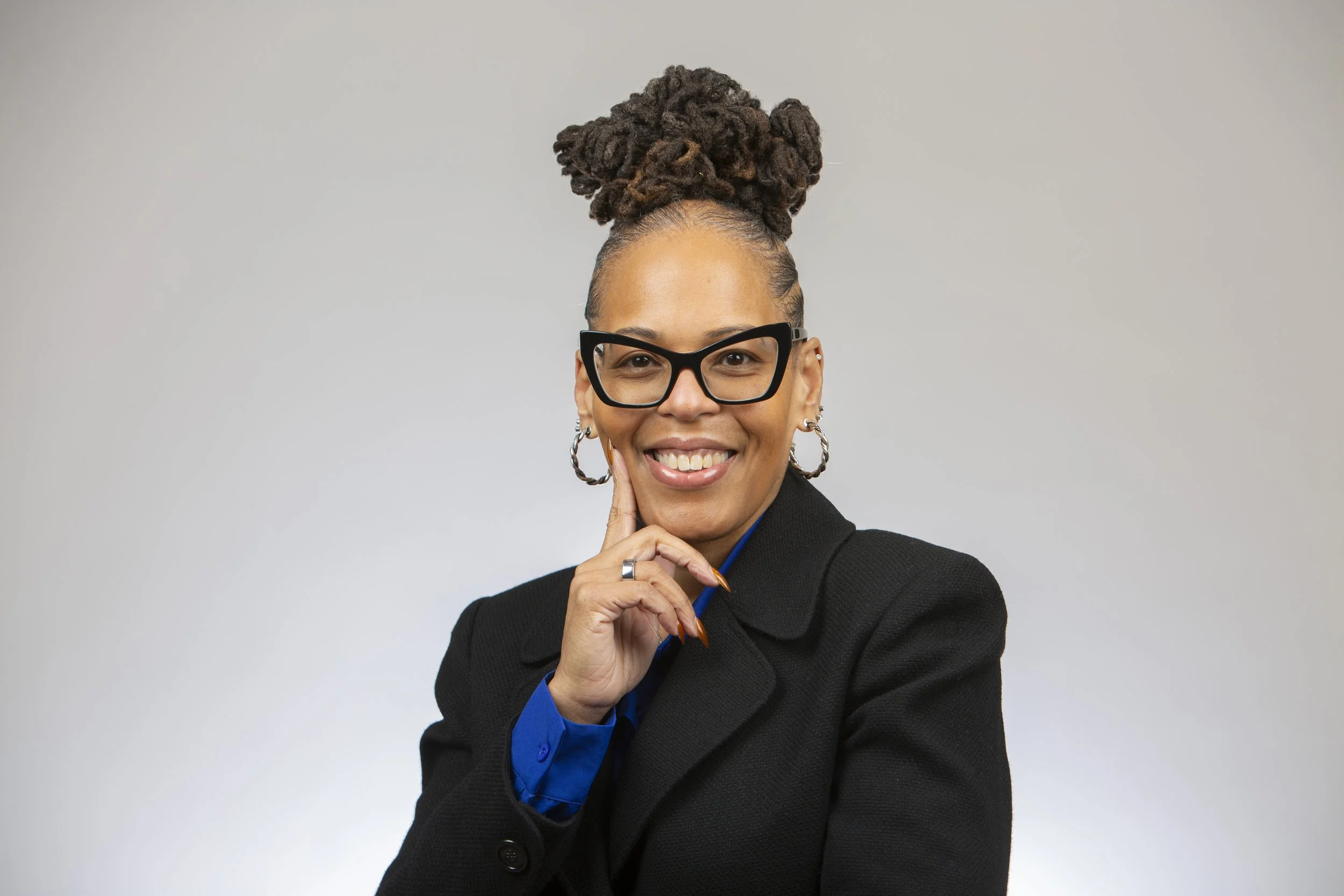 A woman with dark hair styled in dreadlocks hair piled on top of her head, wearing large black glasses, multiple earrings, a black blazer, with a ring, smiling, against a plain light gray background.