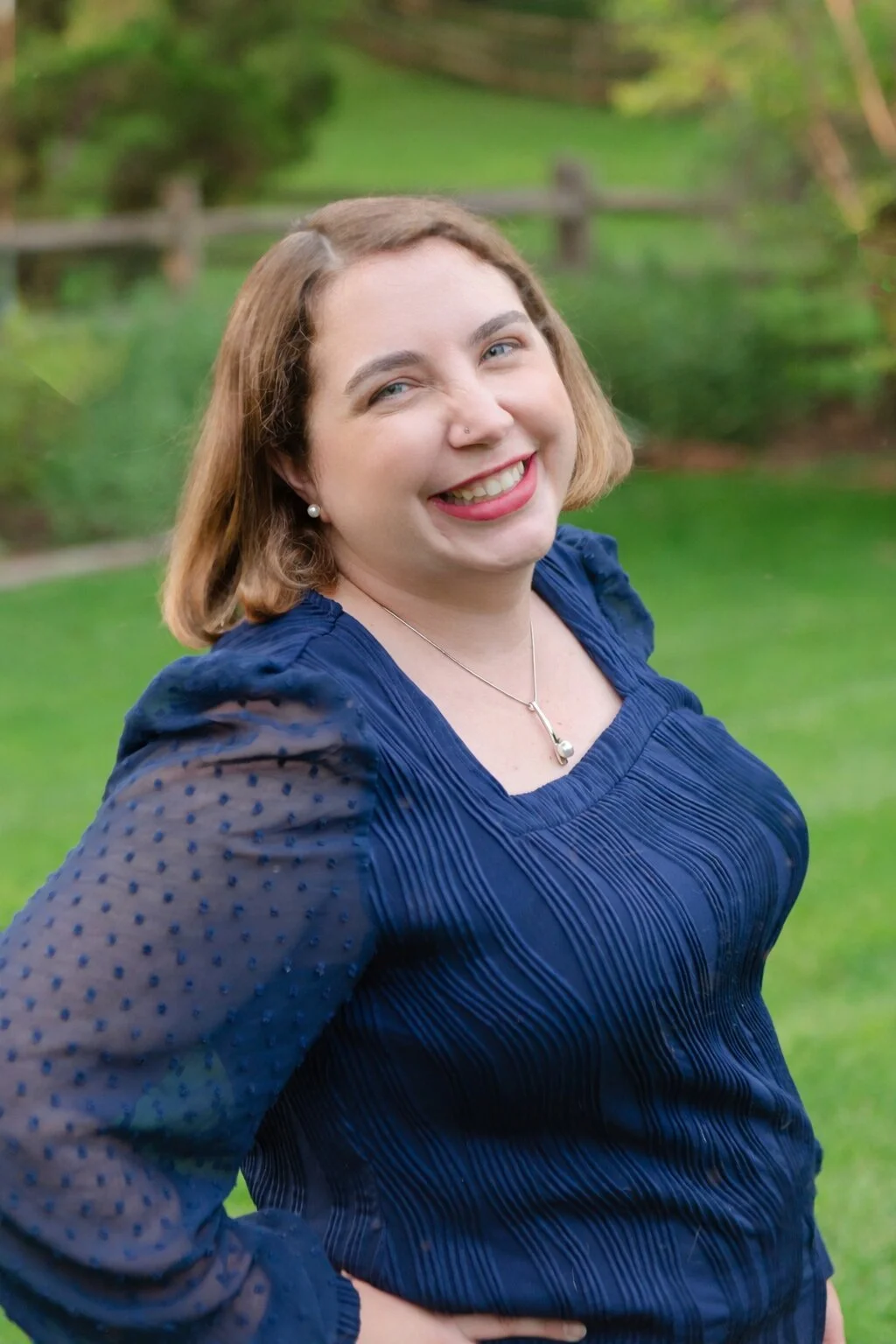 A woman with shoulder-length light brown hair, smiling, wearing a navy blue blouse with sheer polka-dot sleeves, a pearl necklace and earrings, standing outdoors on a grassy area with trees in the background. This is Joanna Miller.