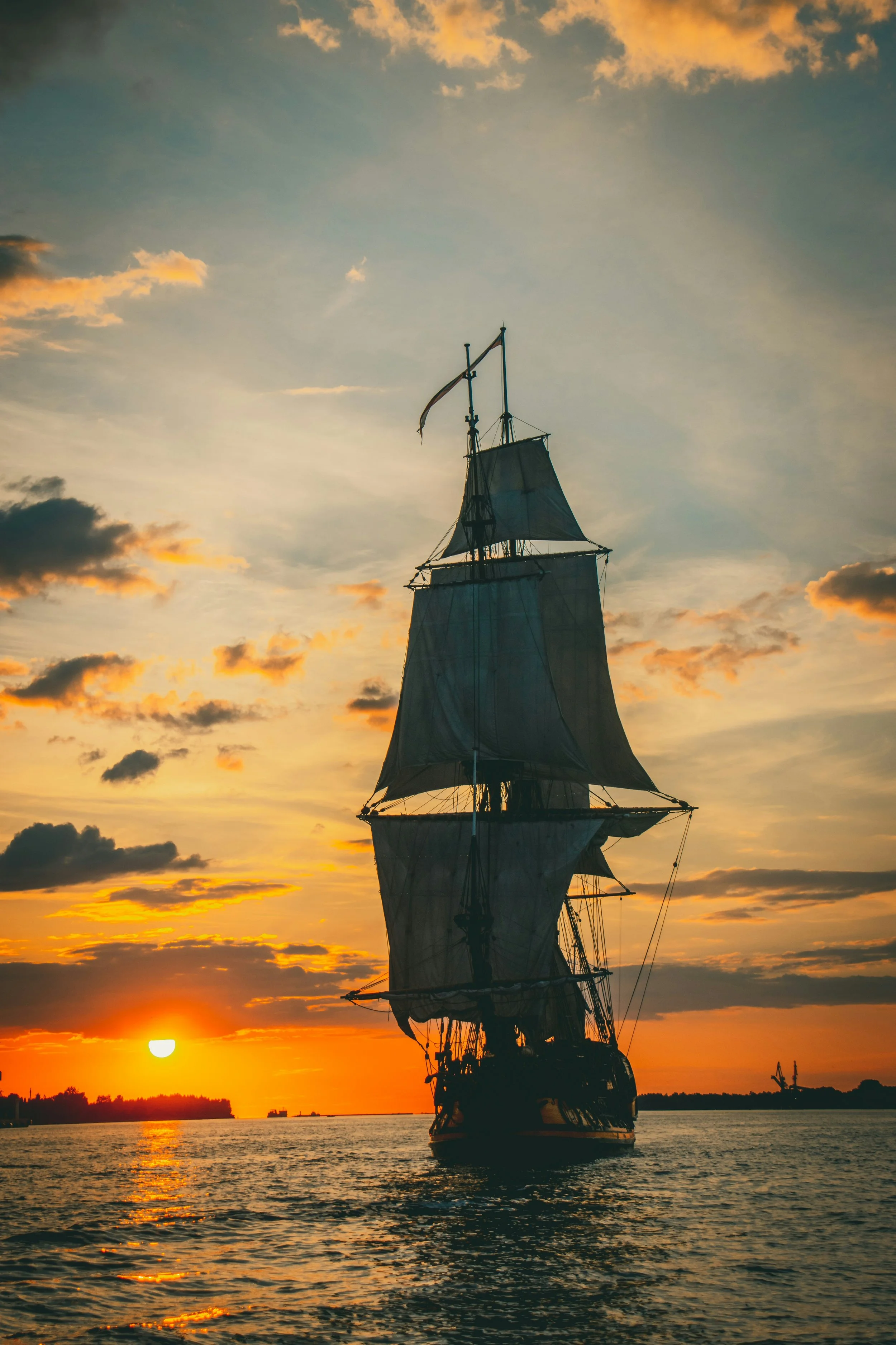A silhouette of a tall ship with sails at sunset, with the sun near the horizon and orange clouds in the sky.