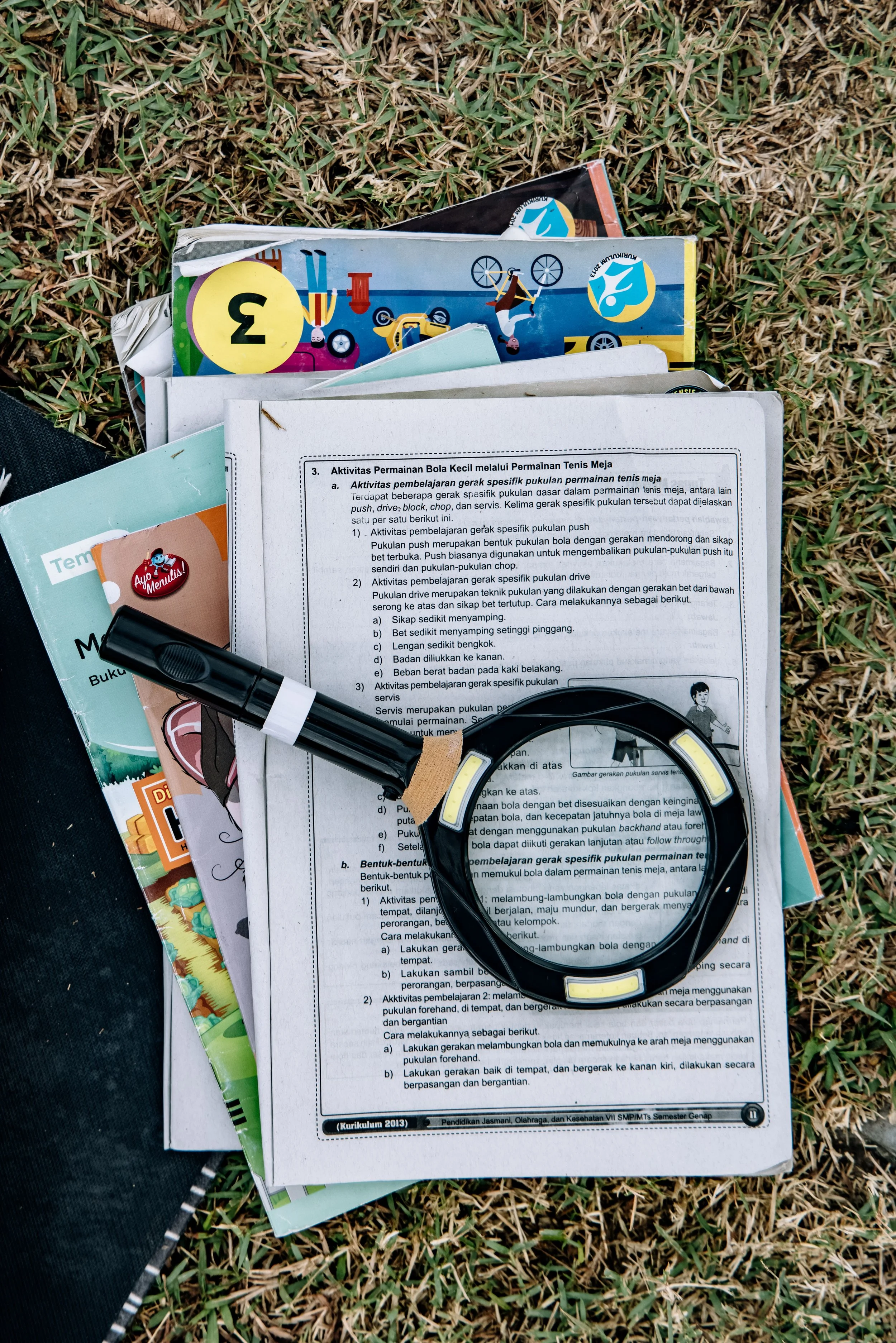 A collection of educational materials, including worksheets, booklets, a magnifying glass, and a marker, scattered on grass, with colorful sports-themed booklets visible among the papers.