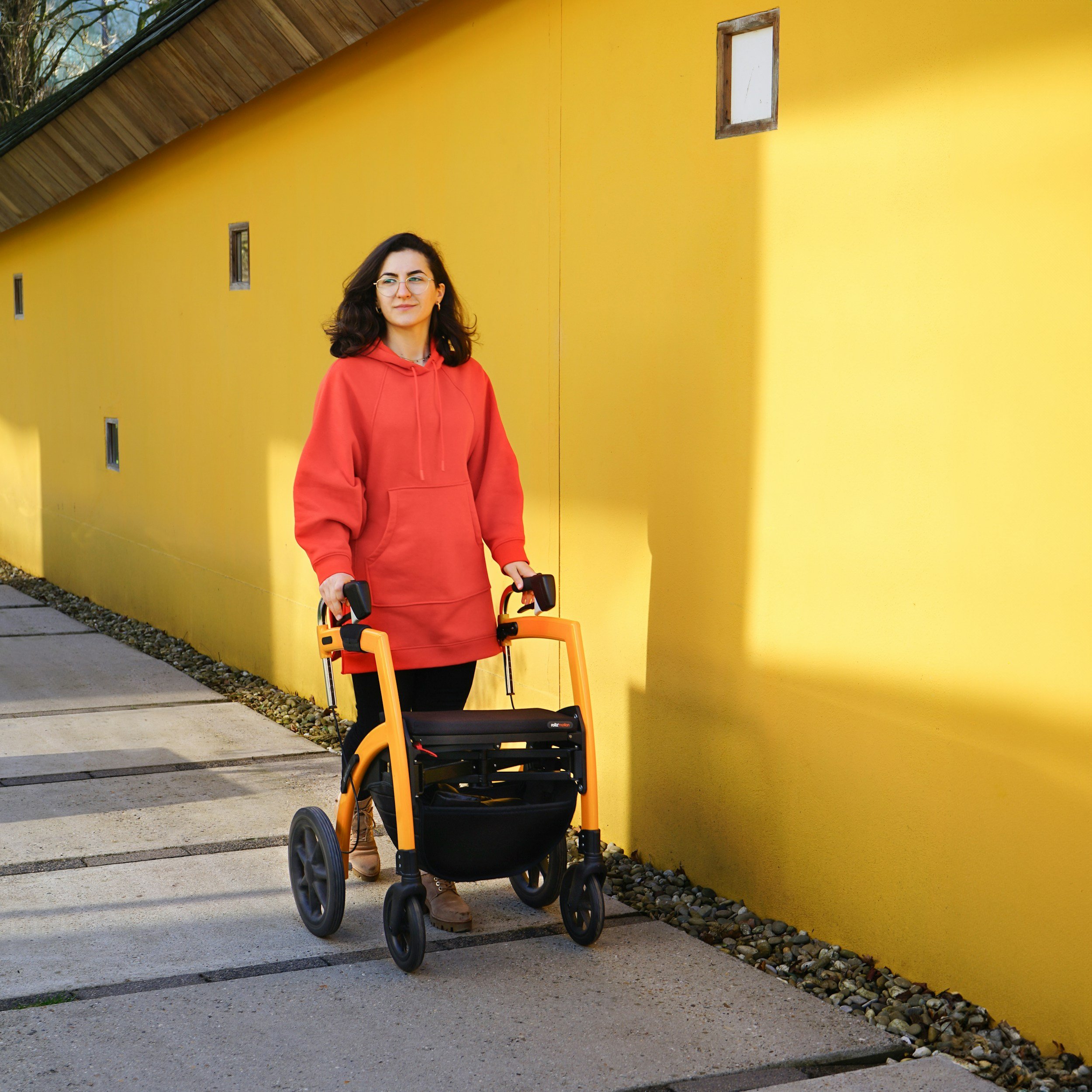 A woman using a walker with dark hair, wearing glasses and a red hoodie, standing outdoors beside a yellow wall with small windows and a sloped roof.