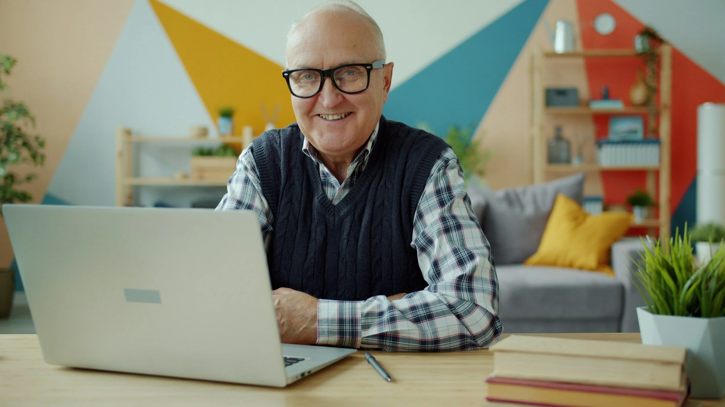 Smiling older man with glasses working on a laptop at a desk in a colorful modern office.
