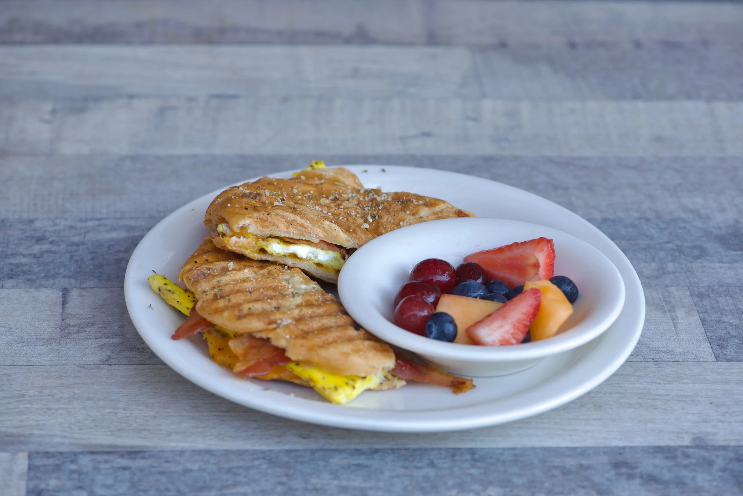 A pressed bacon, egg and cheddar breakfast sandwich cut in half on a white plate, served with a bowl of fresh fruit at Lee's Coffee & Tea in Laurel, Mississippi.