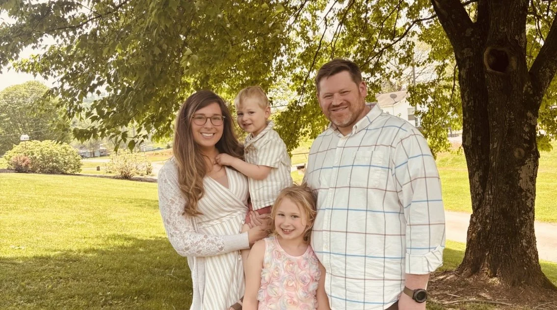 Family of four posing outdoors with trees and a grassy field.