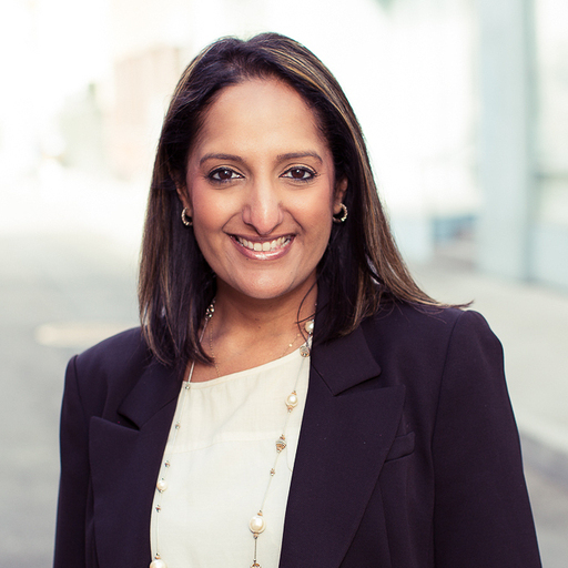 A woman with shoulder-length dark hair and dark eyes smiling, wearing a navy blazer over a white blouse, standing outdoors in an urban setting with modern buildings in the background.