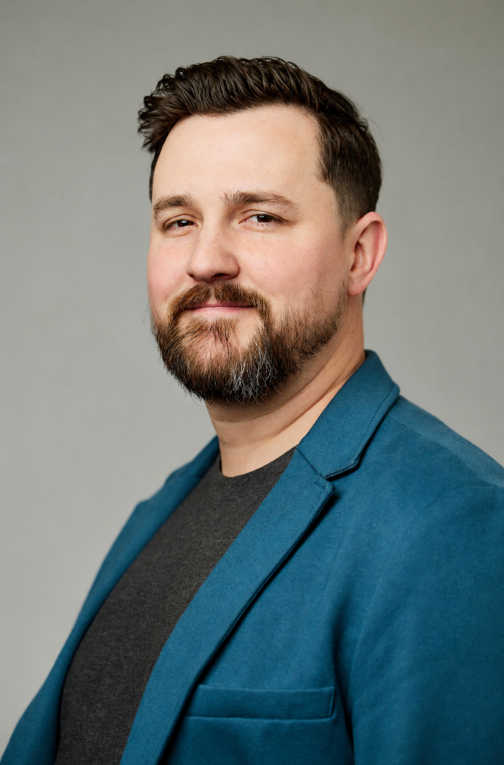 Portrait of a man with brown hair, beard, and mustache, wearing a blue blazer over a dark gray shirt, against a plain gray background.