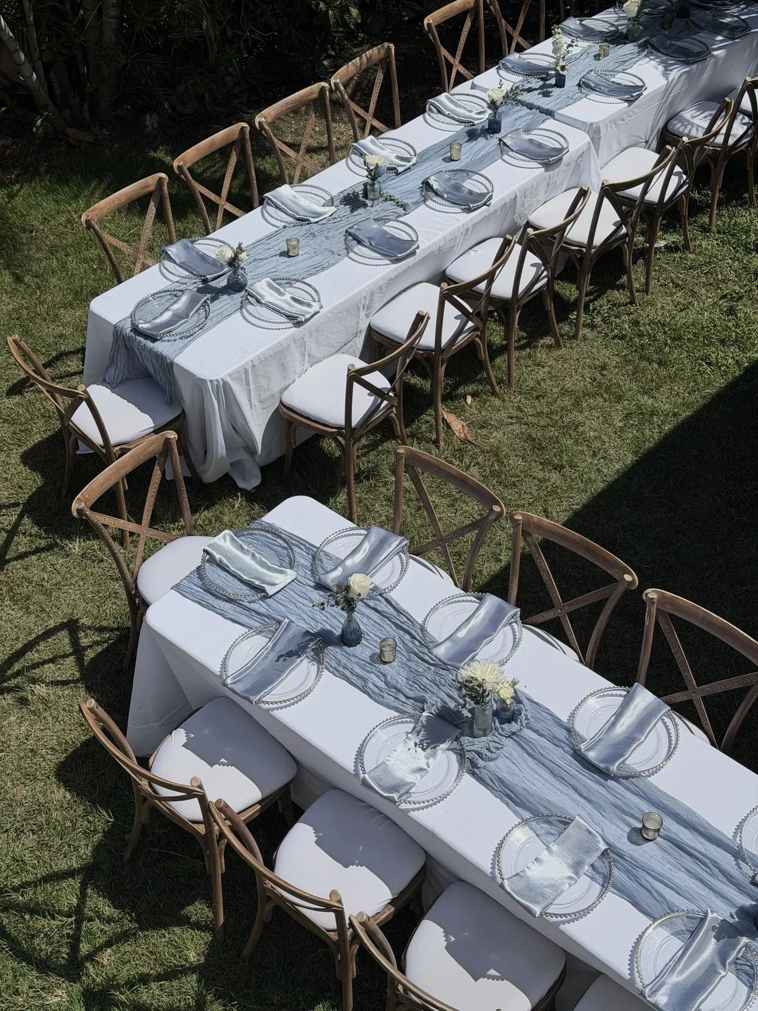 An outdoor dining table setup with white tablecloths, gray and silver tableware, and small floral centerpieces, arranged on a grass lawn with wooden chairs surrounding it.