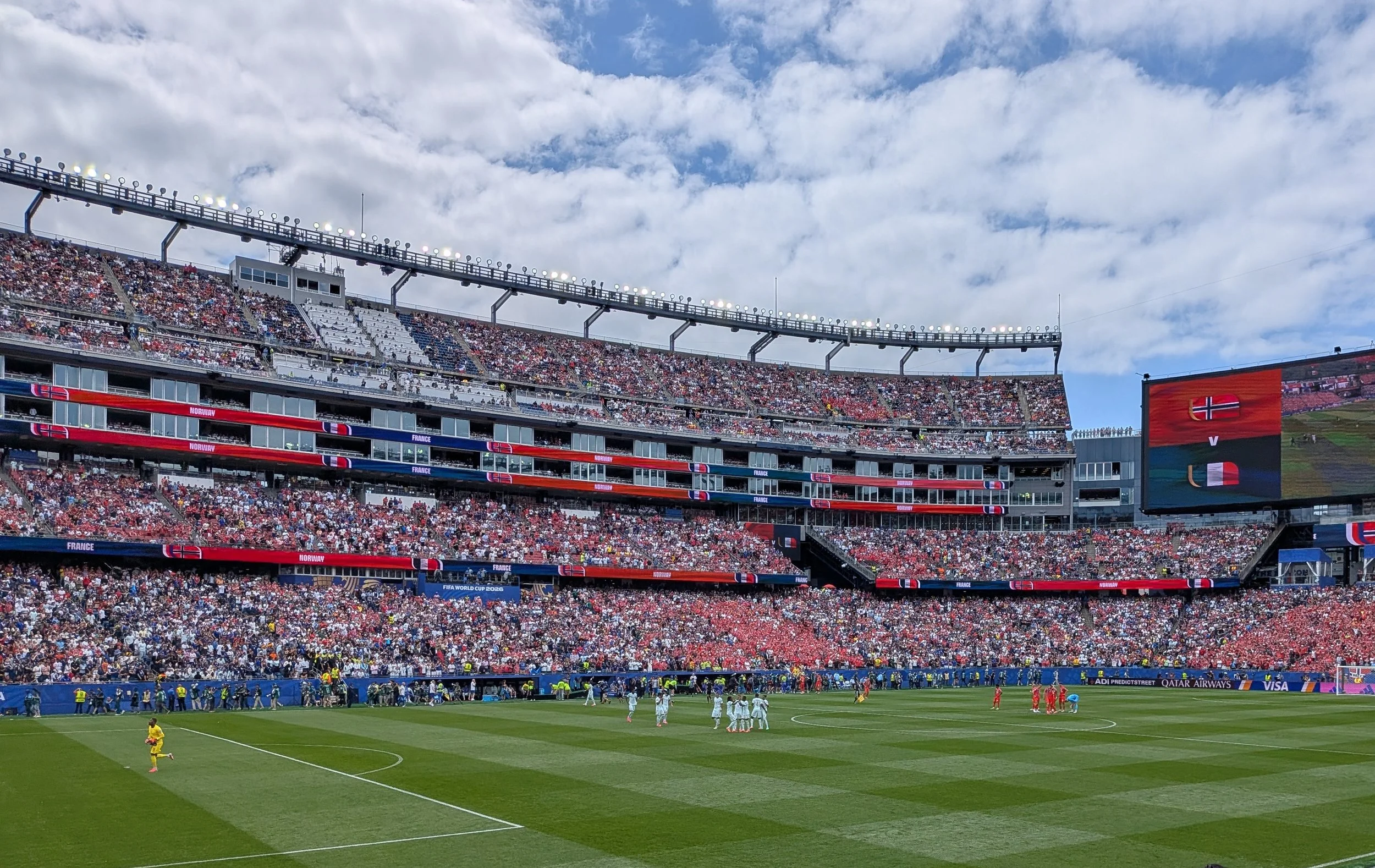 A large stadium filled with spectators during a soccer match. The field is visible at the bottom with players and officials. The stadium has multiple levels with banners displaying Norway and France, indicating an international match, possibly the FIFA World Cup.