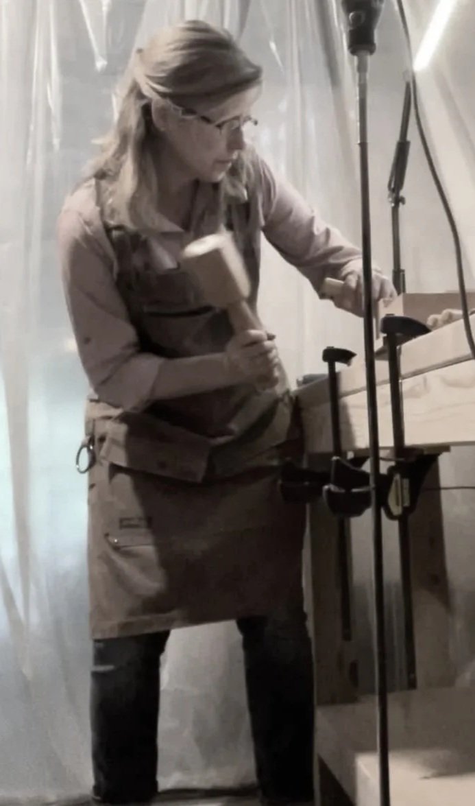 A woman in protective gear is working with a hammer in a workshop, possibly constructing or repairing wooden furniture.