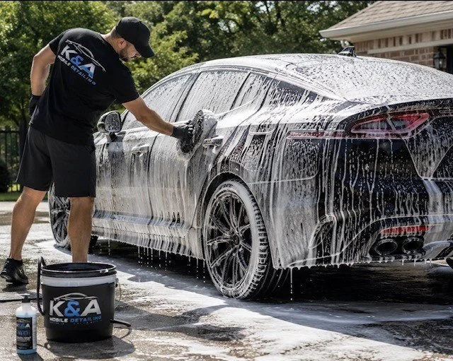 Man washing a black sports car with a foam brush, using soap and water, outdoors in a driveway.