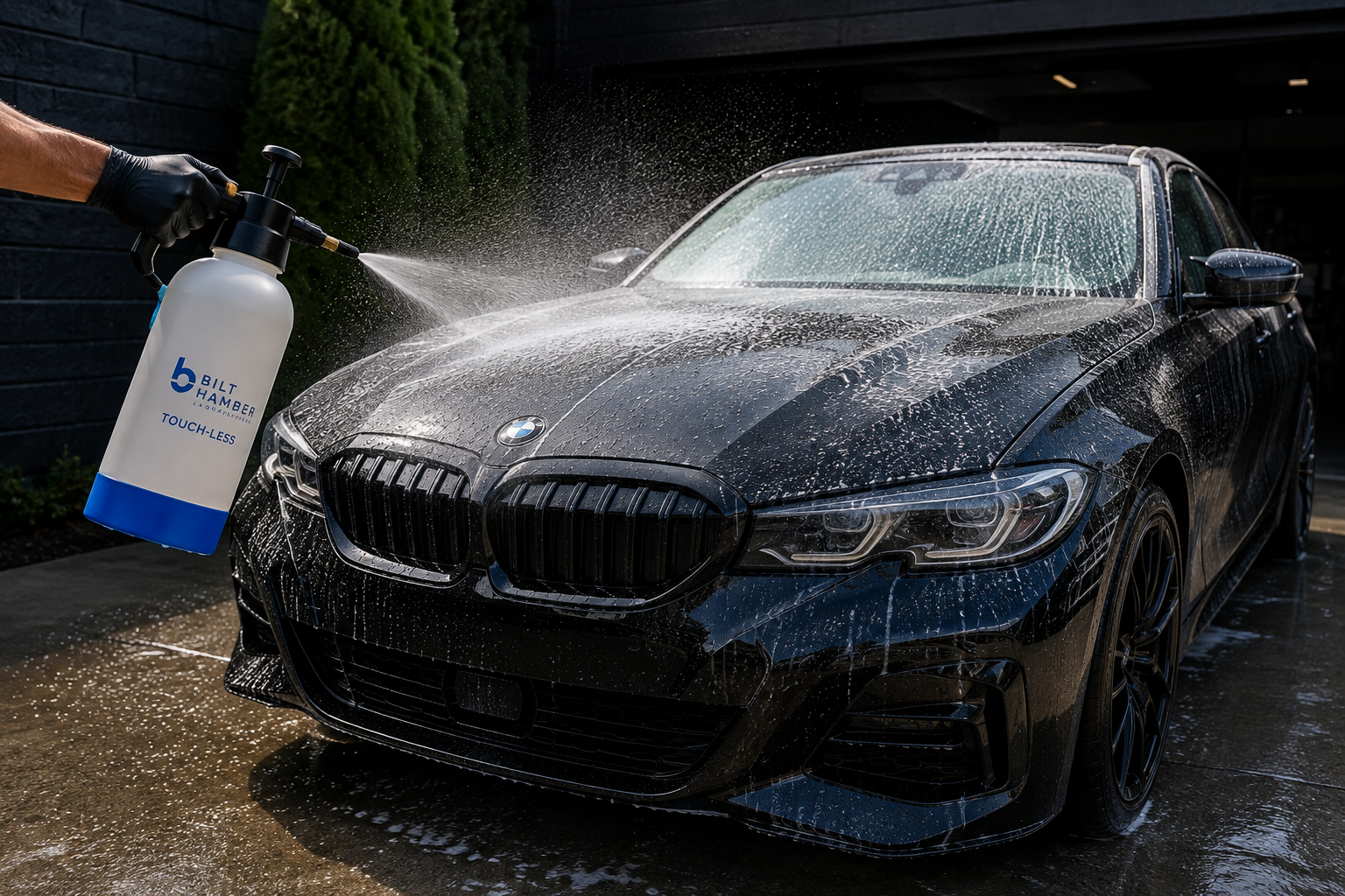 Person wearing black gloves using a spray bottle to wash a black BMW car, with soap suds on the windshield and body, in an outdoor setting.